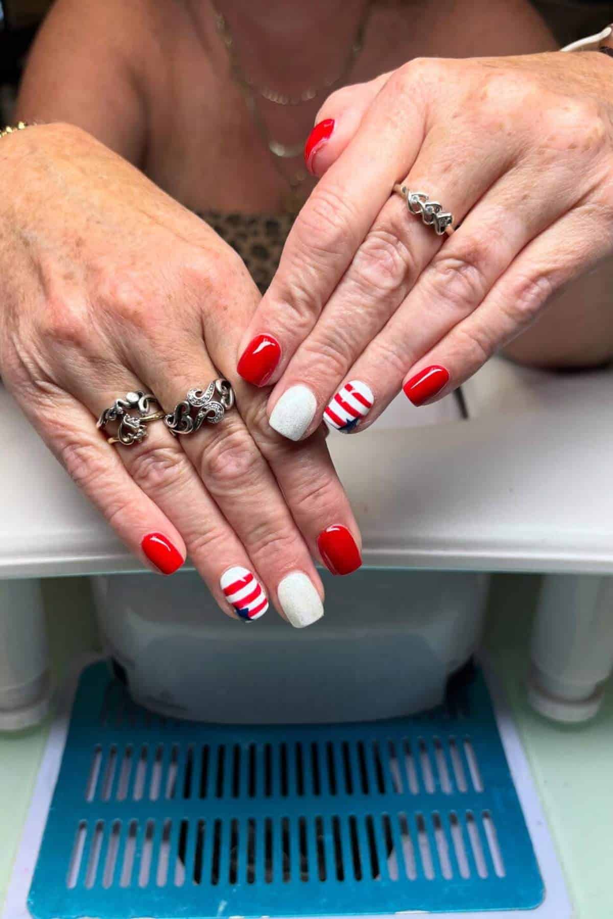 A person displays manicured hands with red, white, and striped nail art; they wear silver rings and rest their hands on a flat surface.