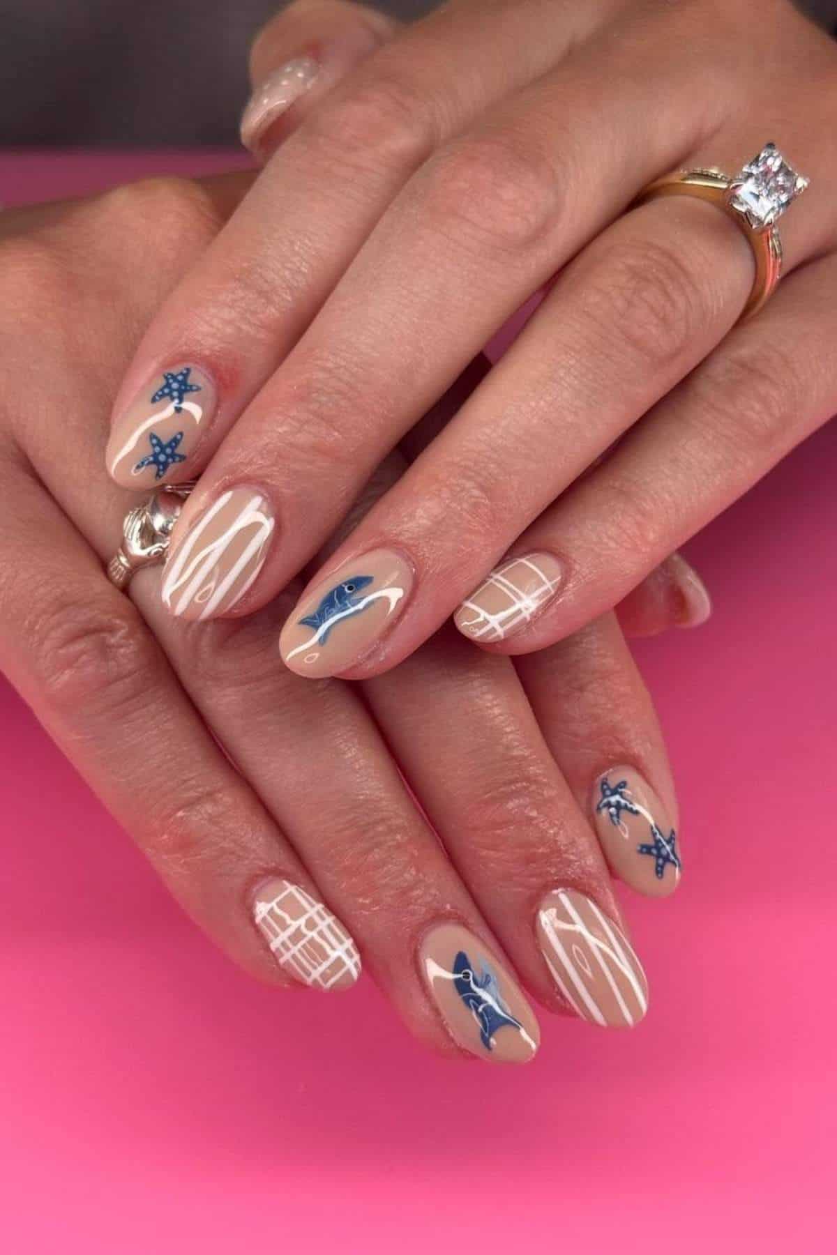 A hand with almond-shaped nails featuring nude polish, blue floral designs, white grid patterns, and abstract white lines, shown against a pink background.