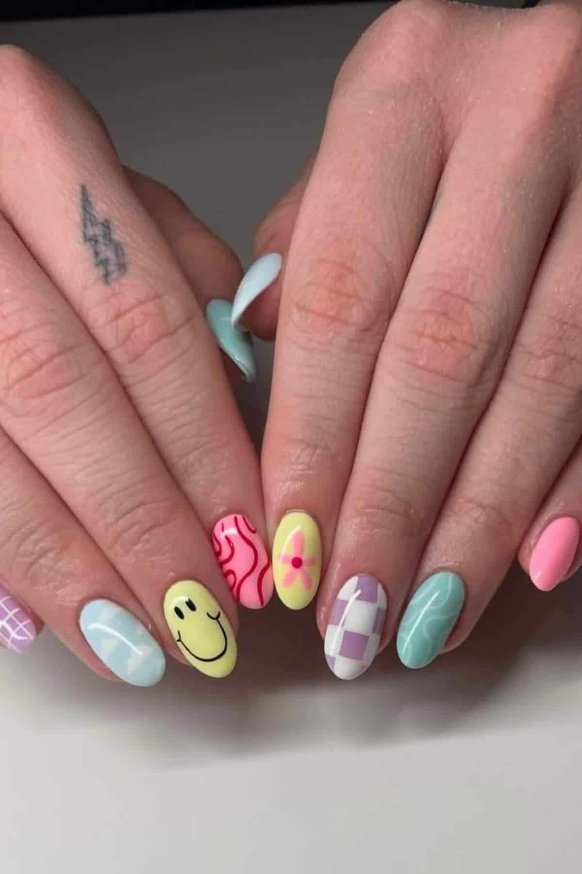 Hands with colorful, patterned nail art including checkerboard, smiley face, flower, and wavy designs; a lightning bolt tattoo is visible on one finger.