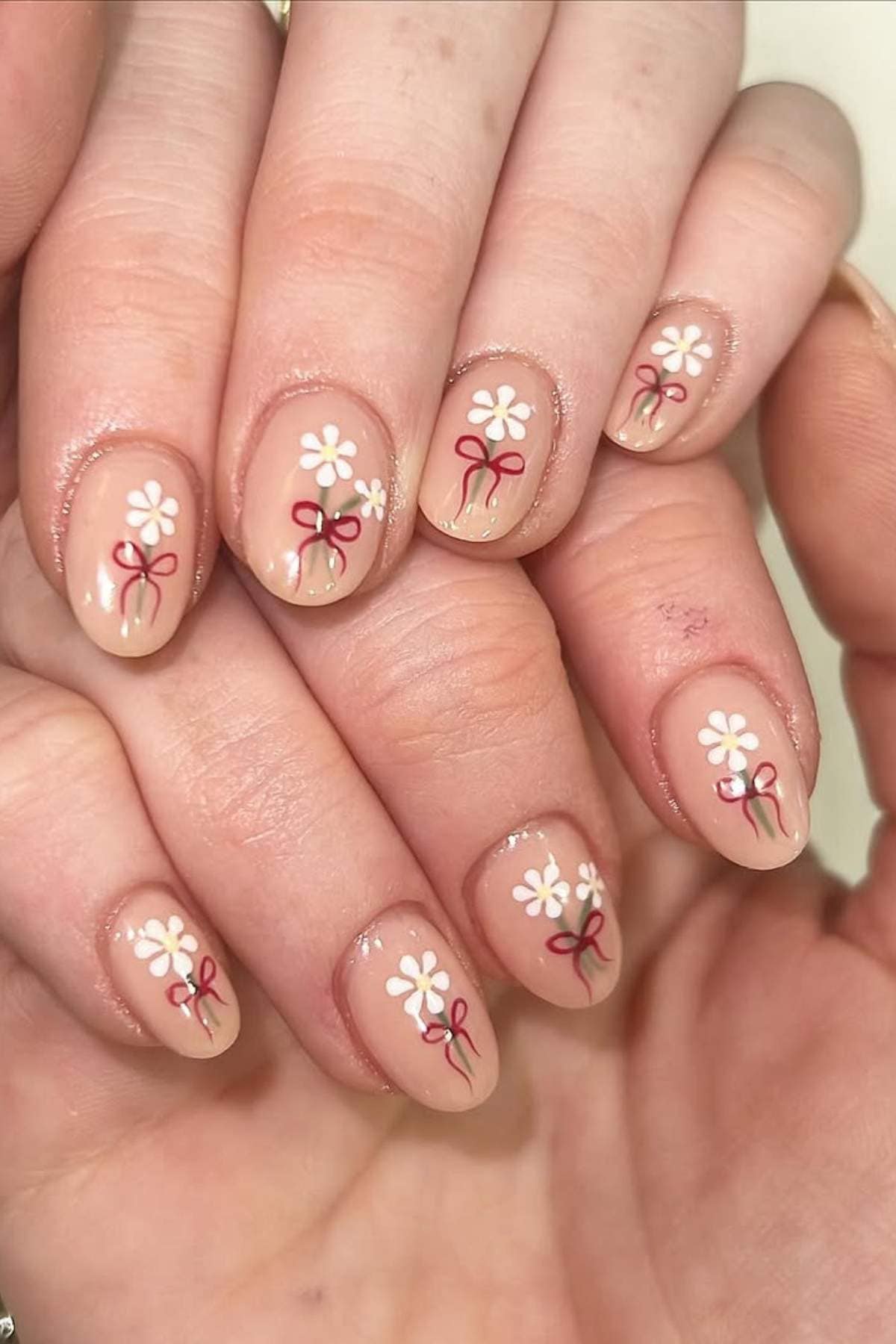 Close-up of hands showing nude-colored nails with small white flower and red bow designs on each nail.