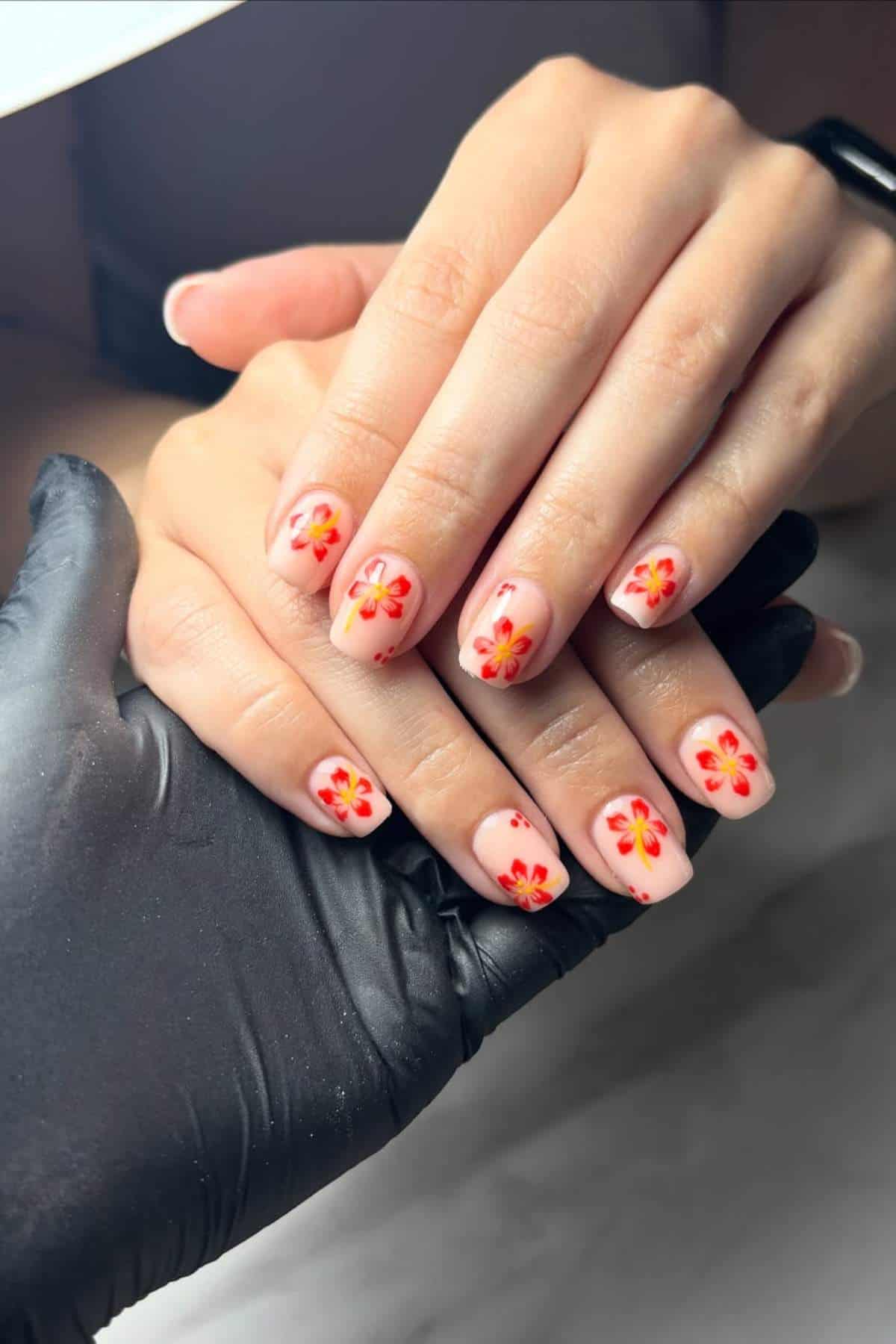 A person displays manicured nails painted with pink polish and decorated with red and yellow floral designs, while another person wearing a black glove holds their hand.
