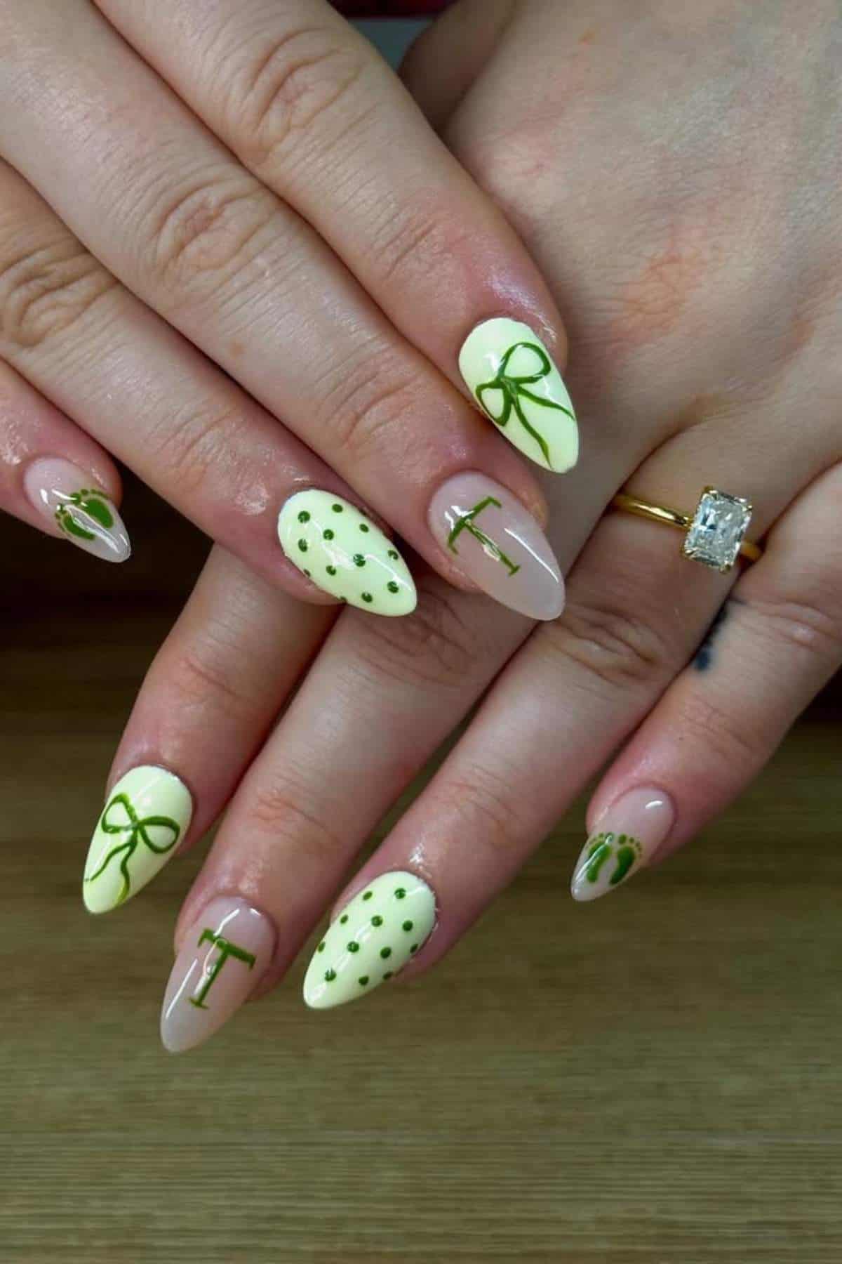 Hands with long almond-shaped nails featuring green polka dots, bows, and the letters "I" and "T" on a pale yellow and nude base. Person wears a gold ring with a large rectangular stone.