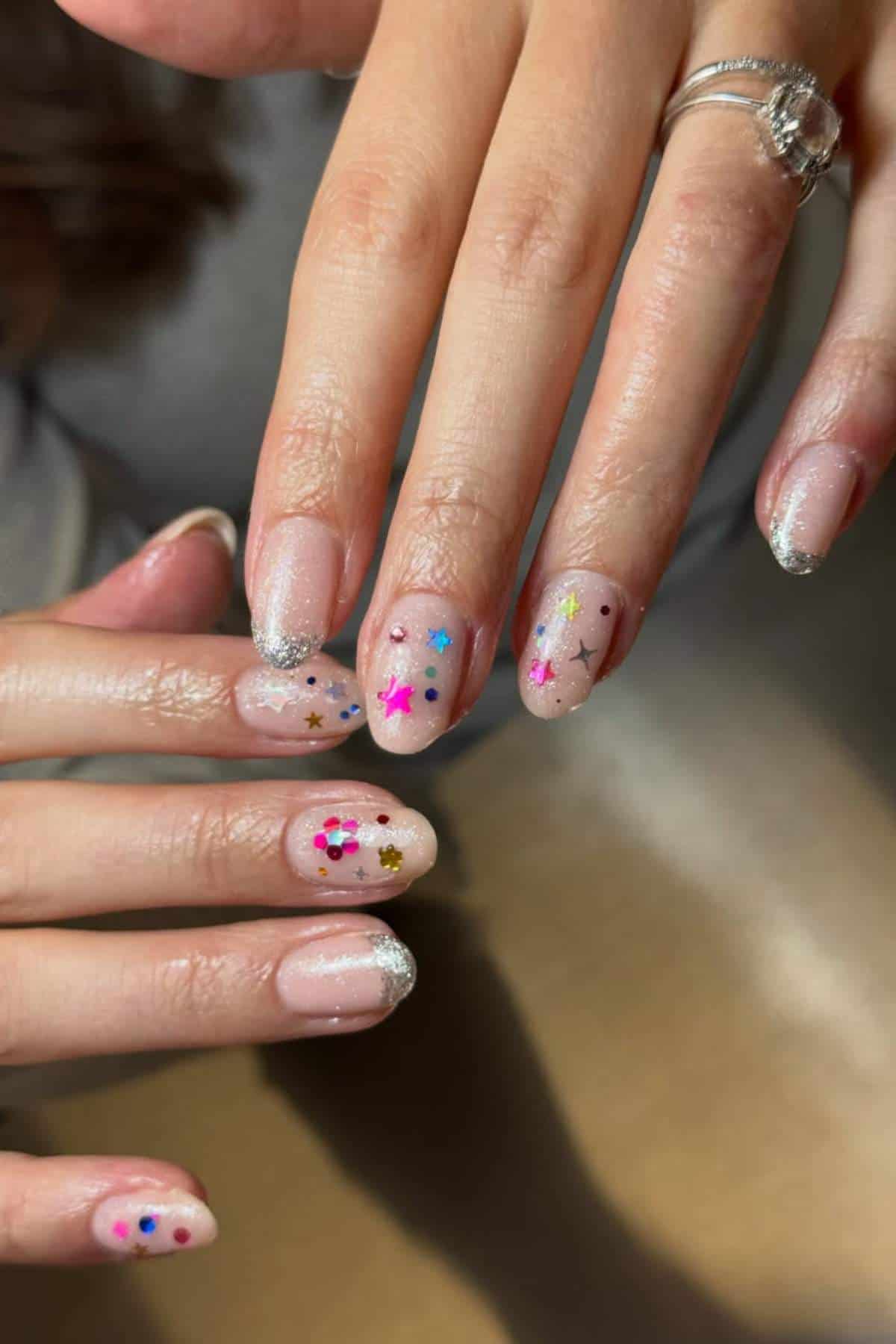 Close-up of hands with light pink manicure, silver glitter tips, and colorful star and dot nail art; person is wearing a ring with a large gem.