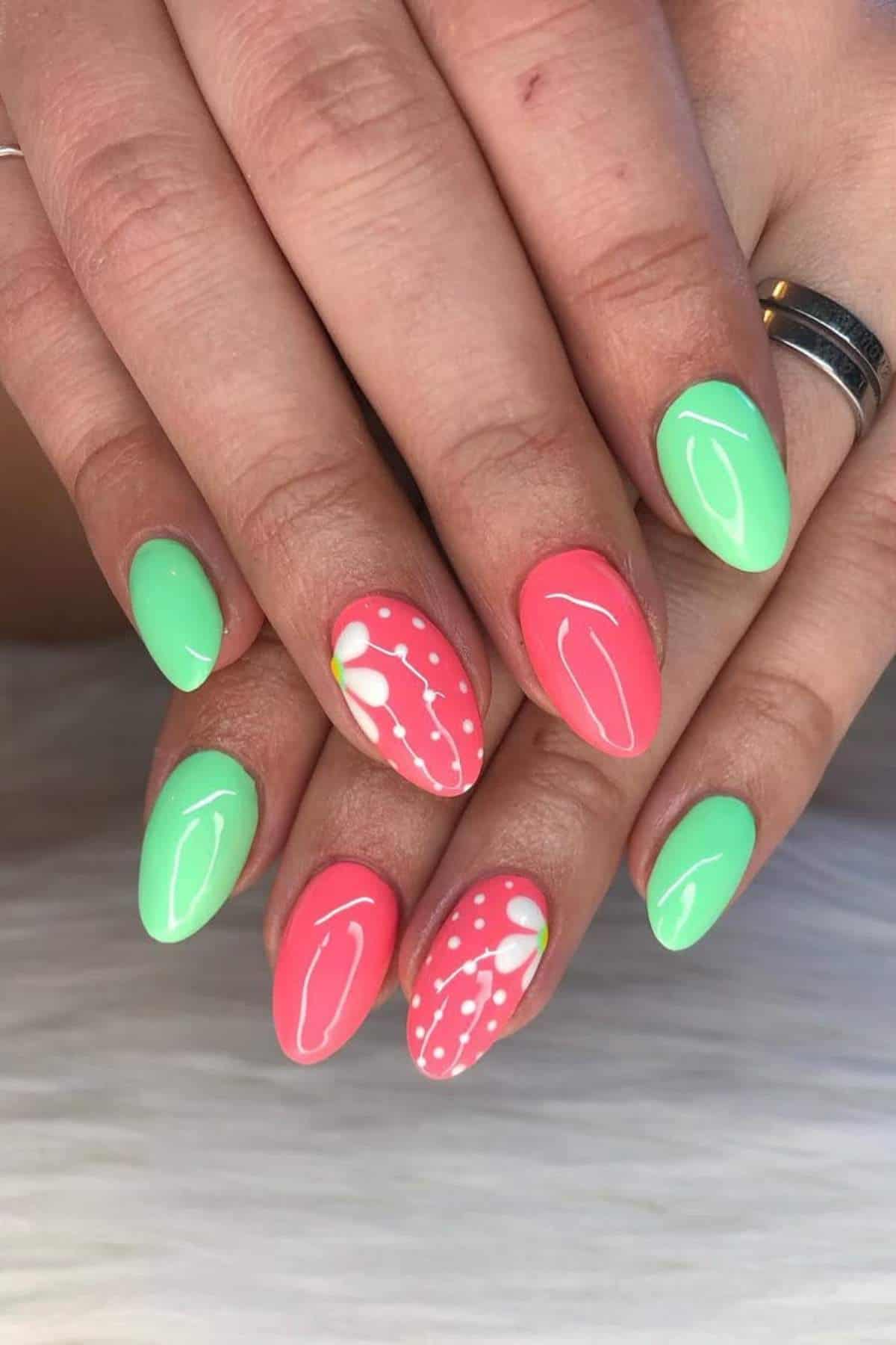 Hands with almond-shaped nails painted in alternating mint green and coral pink, with white floral and dotted designs on two pink nails.