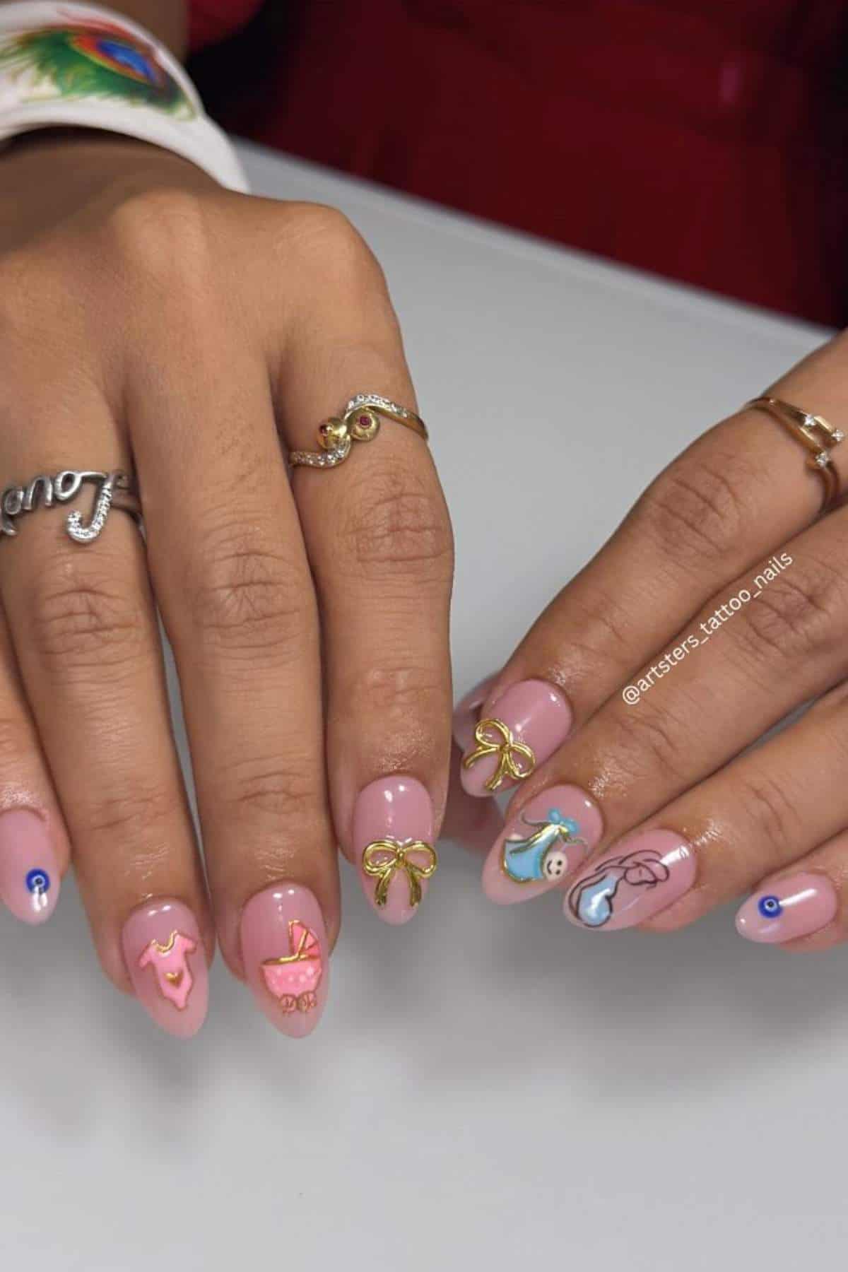 Hands with pink manicured nails featuring 3D gold bows, evil eye decals, and other decorative nail art; person is wearing gold rings.