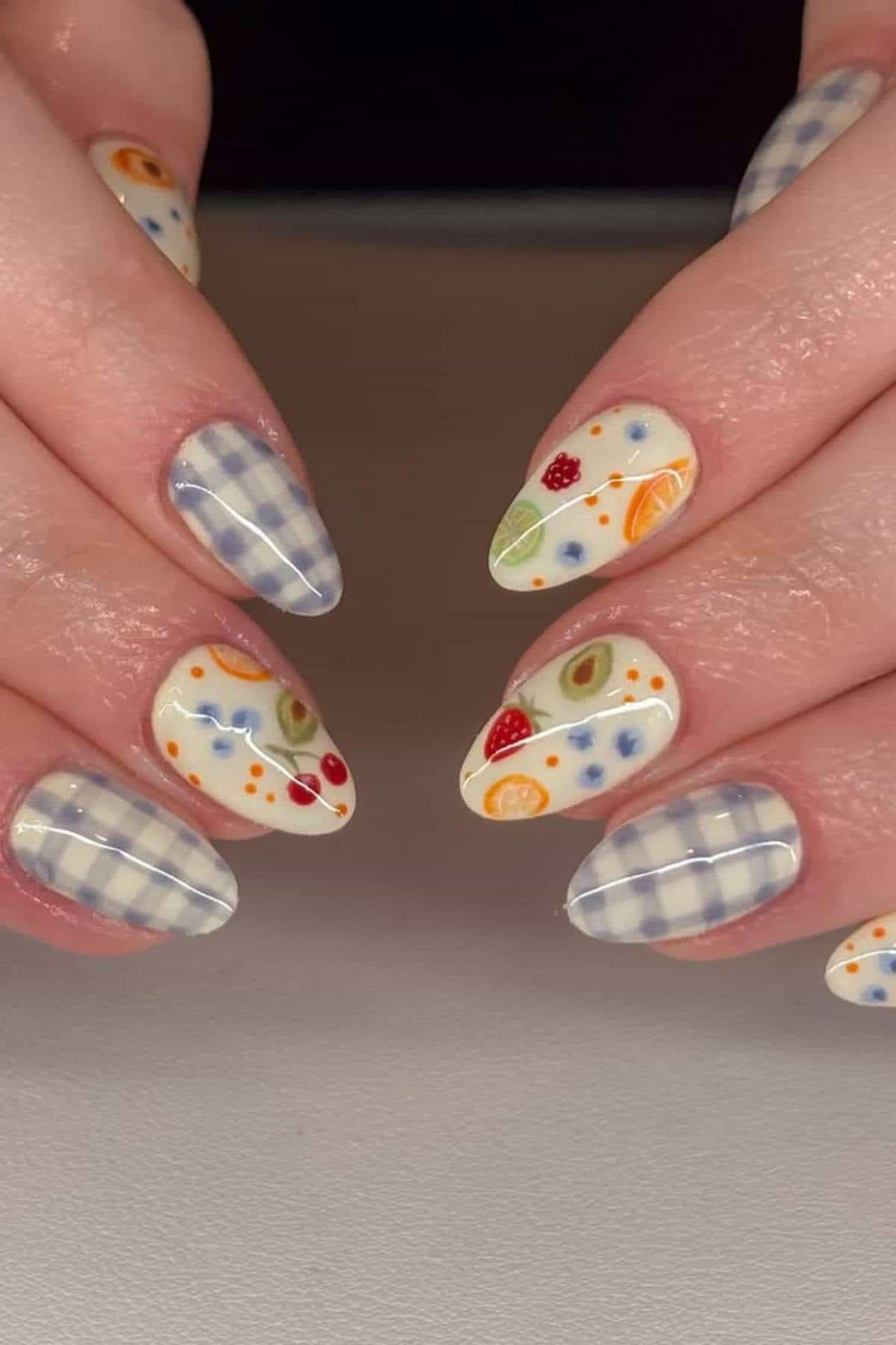 Close-up of hands with almond-shaped nails featuring blue and white plaid patterns on some nails and colorful fruit designs on others.