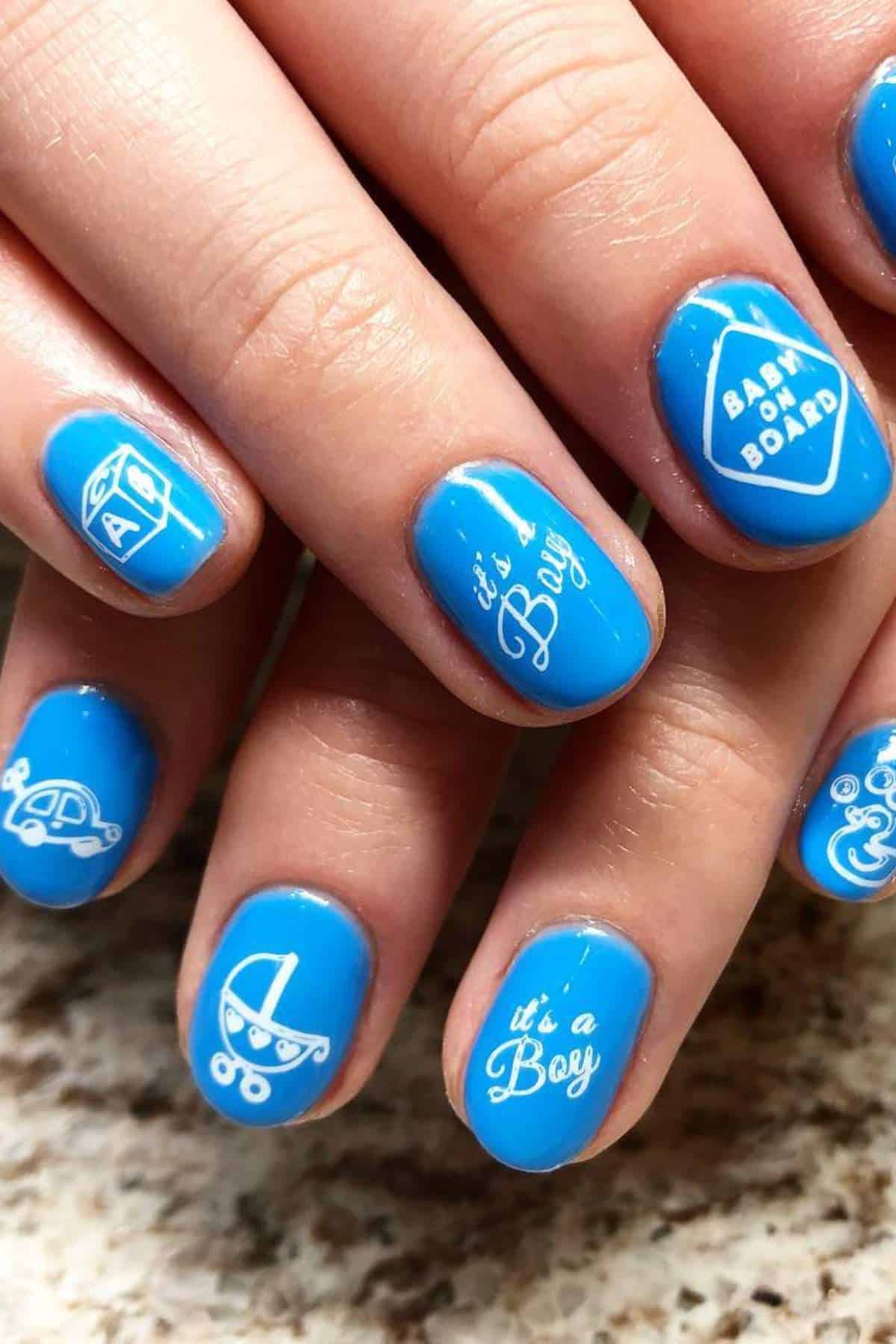 Hands with short, rounded nails painted blue, featuring white baby-themed designs and text such as "It's a Boy," "Baby on Board," a stroller, bottle, and car.