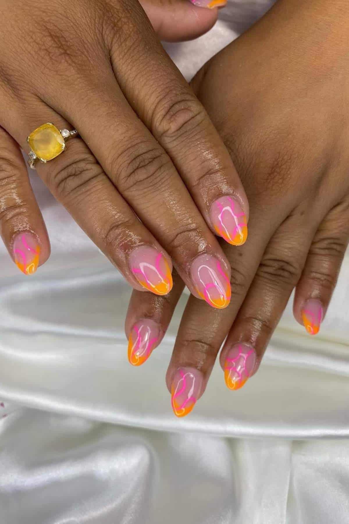 Hands with almond-shaped nails painted with a pink and orange neon swirl design, resting on white fabric. One hand wears a yellow gemstone ring.