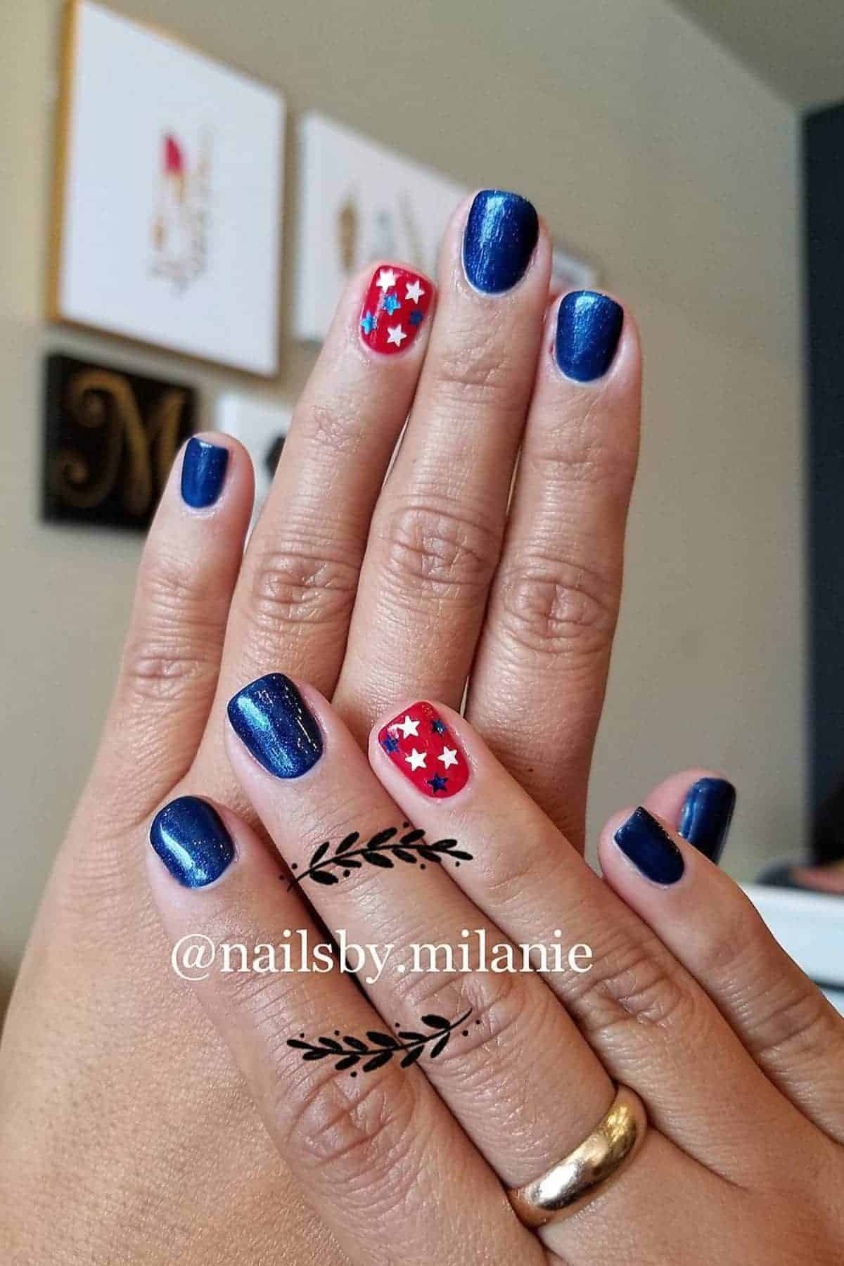 Hands with short, dark blue nails; ring fingers feature a red base with white stars and blue dots. Gold ring on one finger. "@nailsby.milanie" watermark on image.