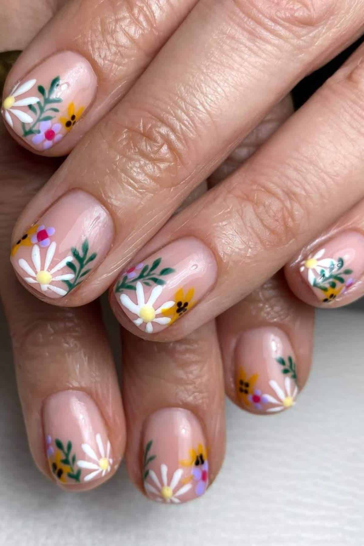Close-up of fingernails painted with a clear base and decorated with colorful floral designs, including white, yellow, and pink flowers with green leaves.