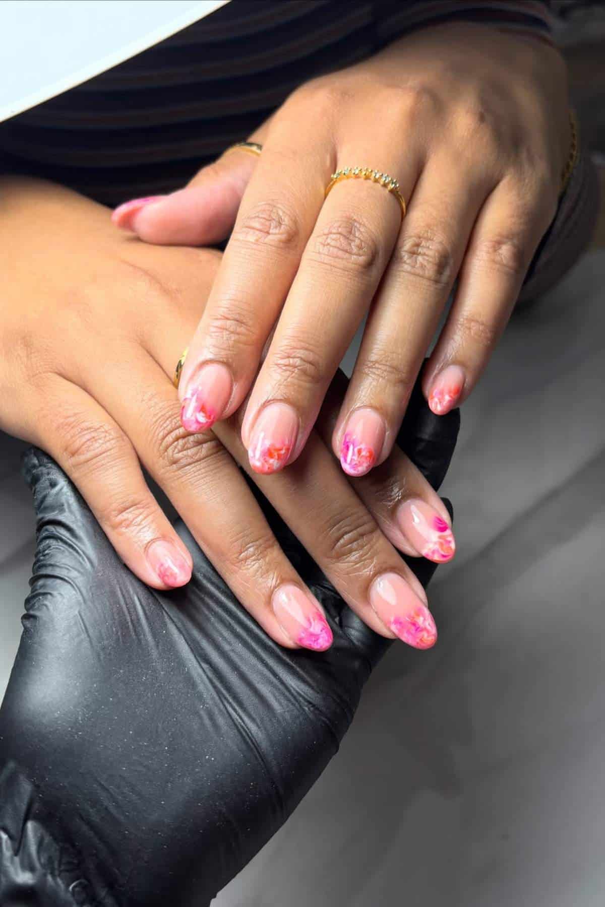 A person with almond-shaped nails featuring a pink and nude gradient design with floral accents, hands resting on a surface, one gloved hand supporting.