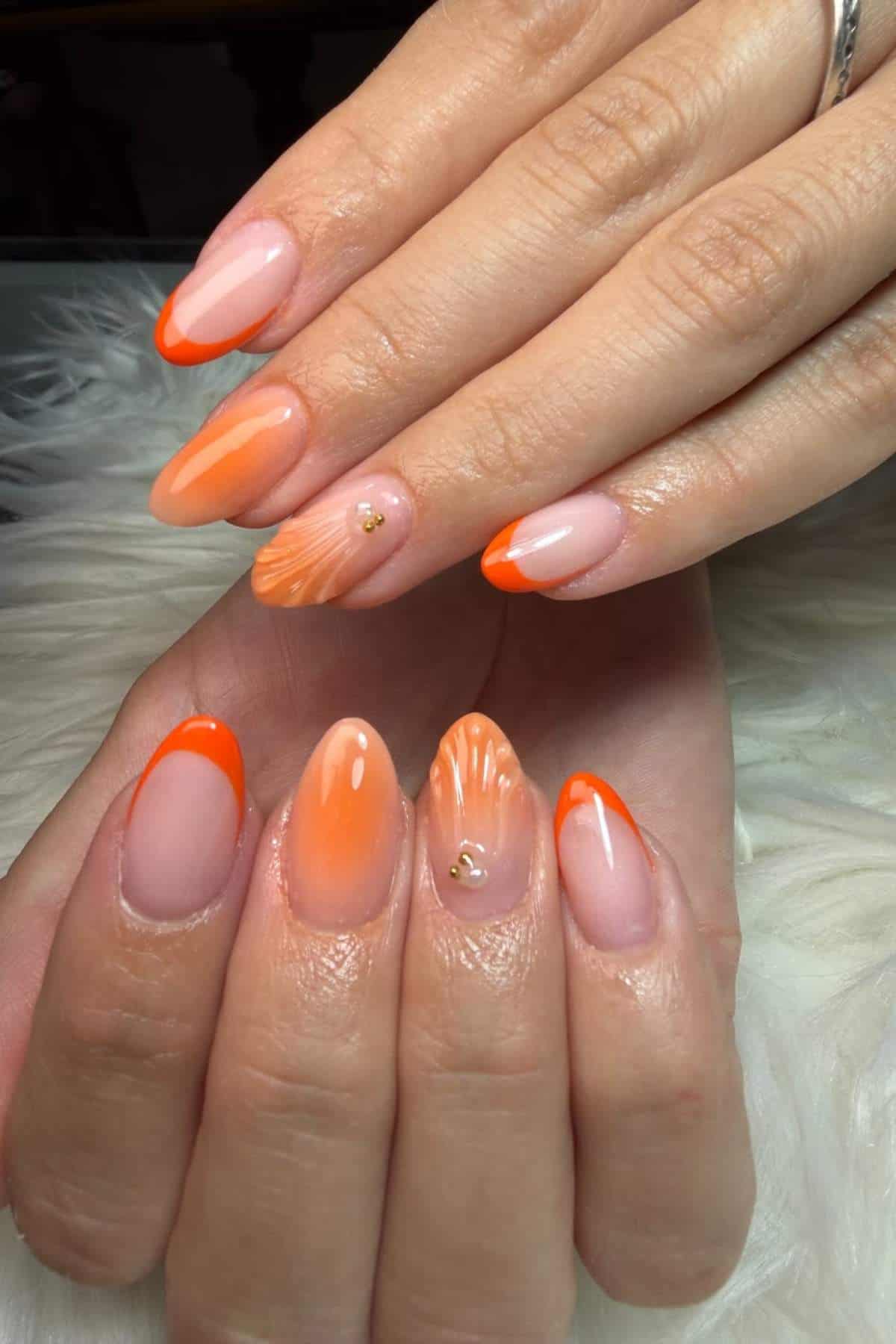 Hands with almond-shaped nails featuring an orange French tip design, abstract orange patterns, and small gold bead accents on two nails.