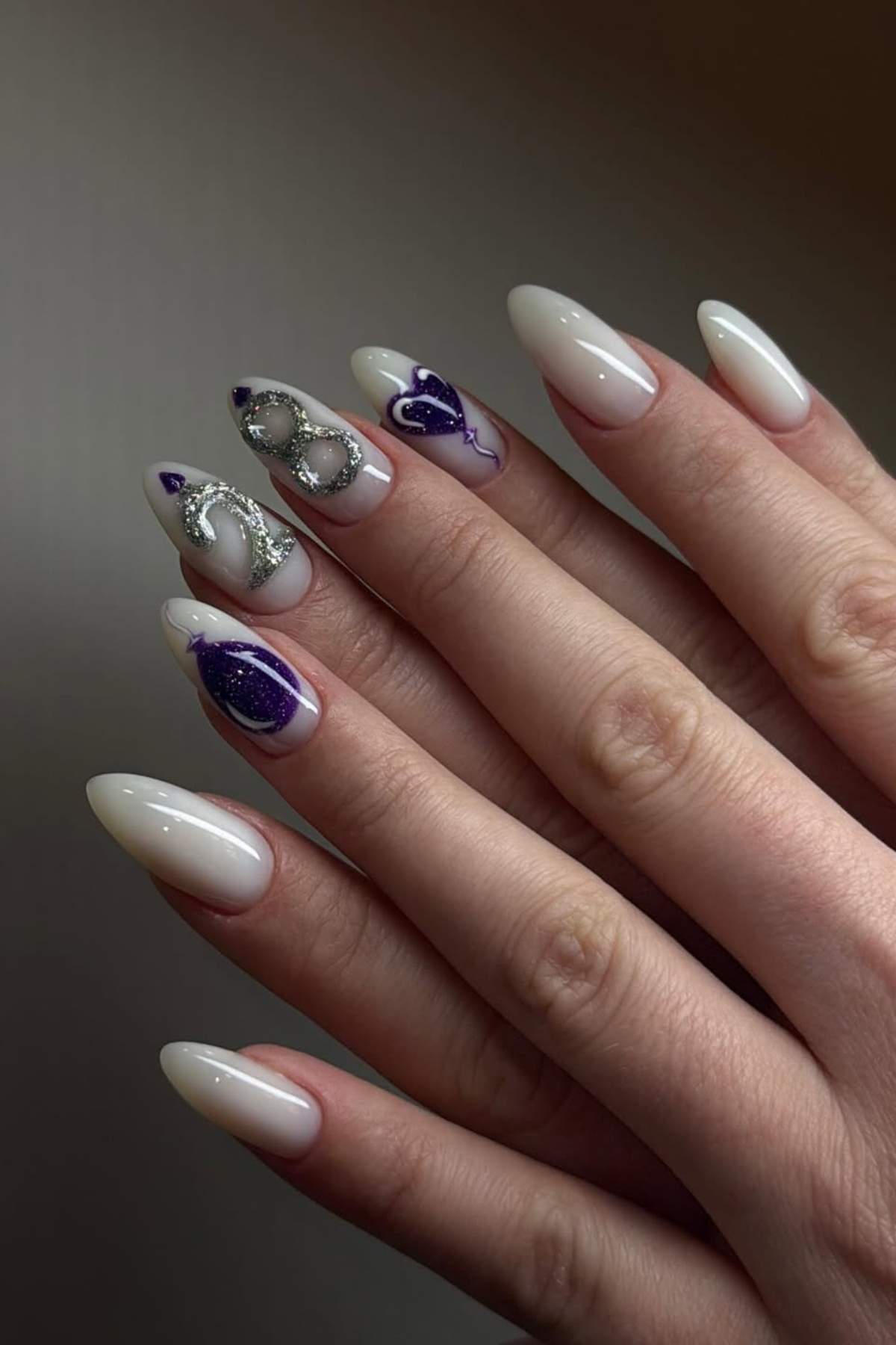 A hand with almond-shaped nails painted in a nude polish, featuring purple and silver abstract designs on the middle and ring fingers.