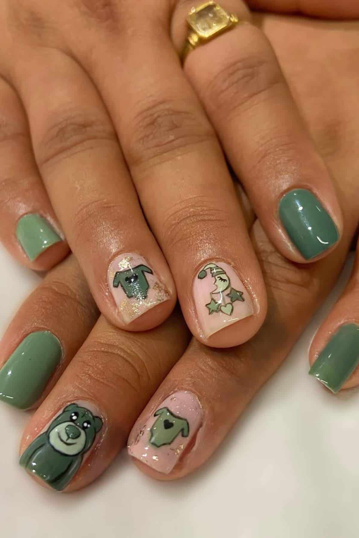 Close-up of hands with green and nude nails featuring hand-painted designs of a bear, T-shirts, and a heart with holly accents. Person is wearing a gold ring.