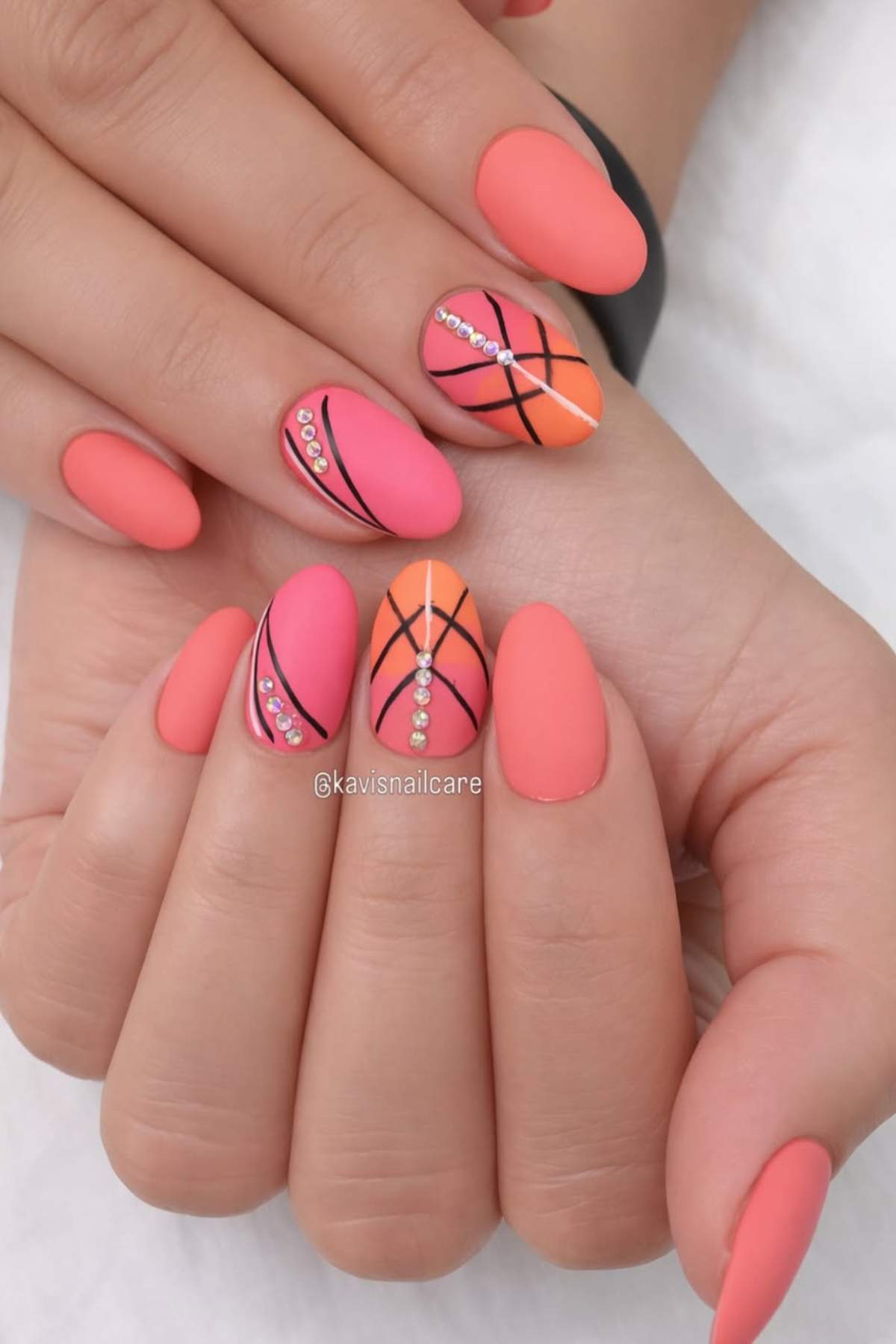 Hands with oval-shaped nails painted in shades of coral and orange, featuring black lines, rhinestone accents, and matte finish.