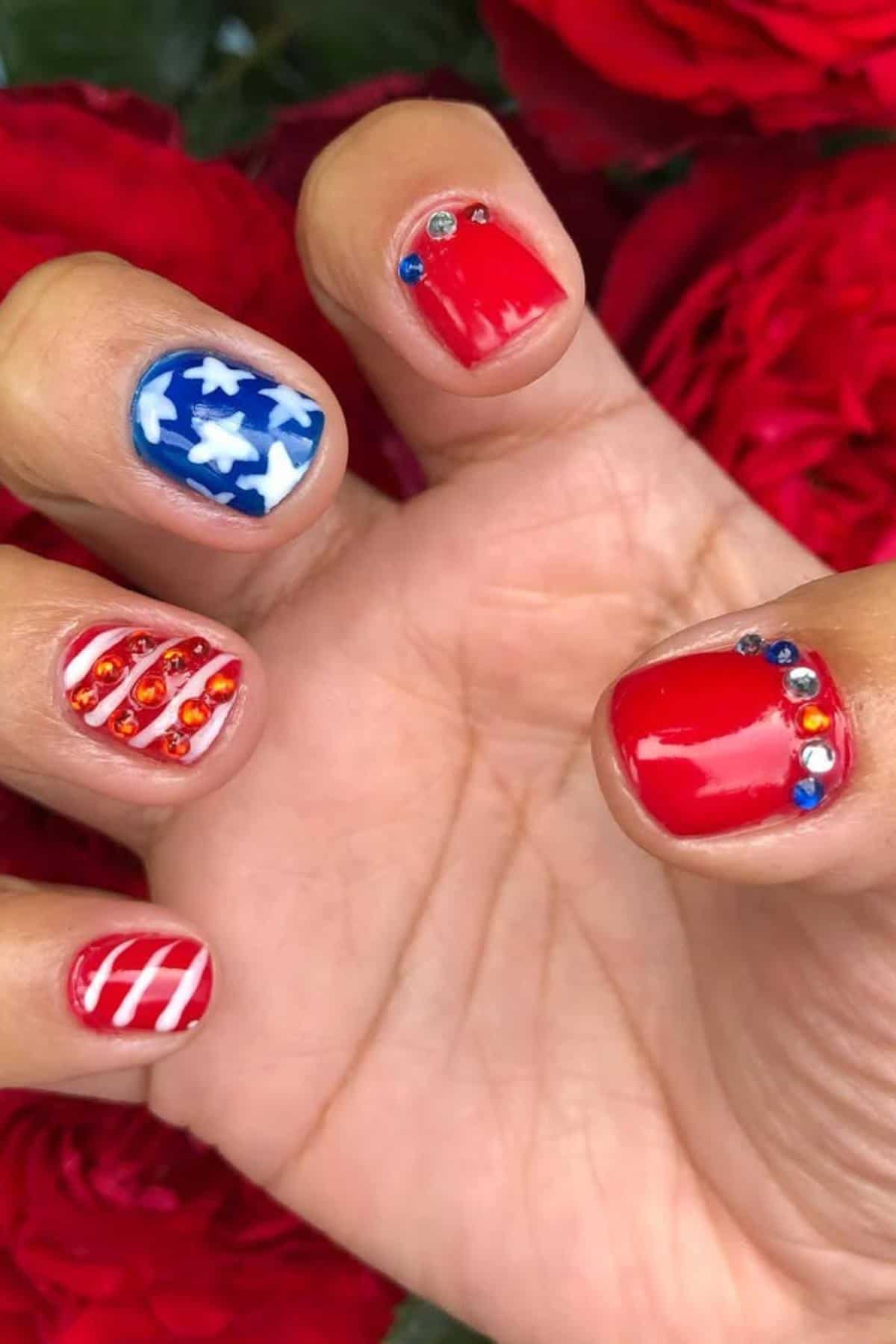 A hand with fingernails painted in red, white, and blue designs, decorated with rhinestones and patterns resembling the American flag. Red roses are in the background.