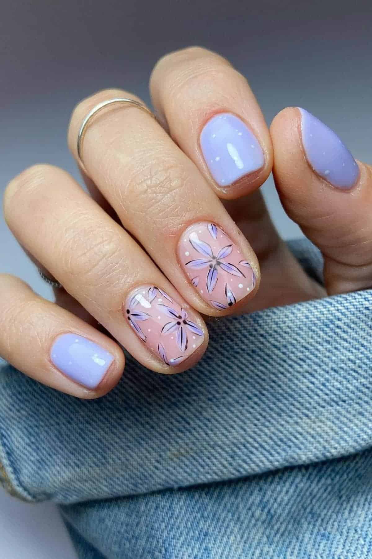 Hand with short nails painted light purple and nude pink; two nails feature purple floral designs. The hand rests on blue denim fabric.