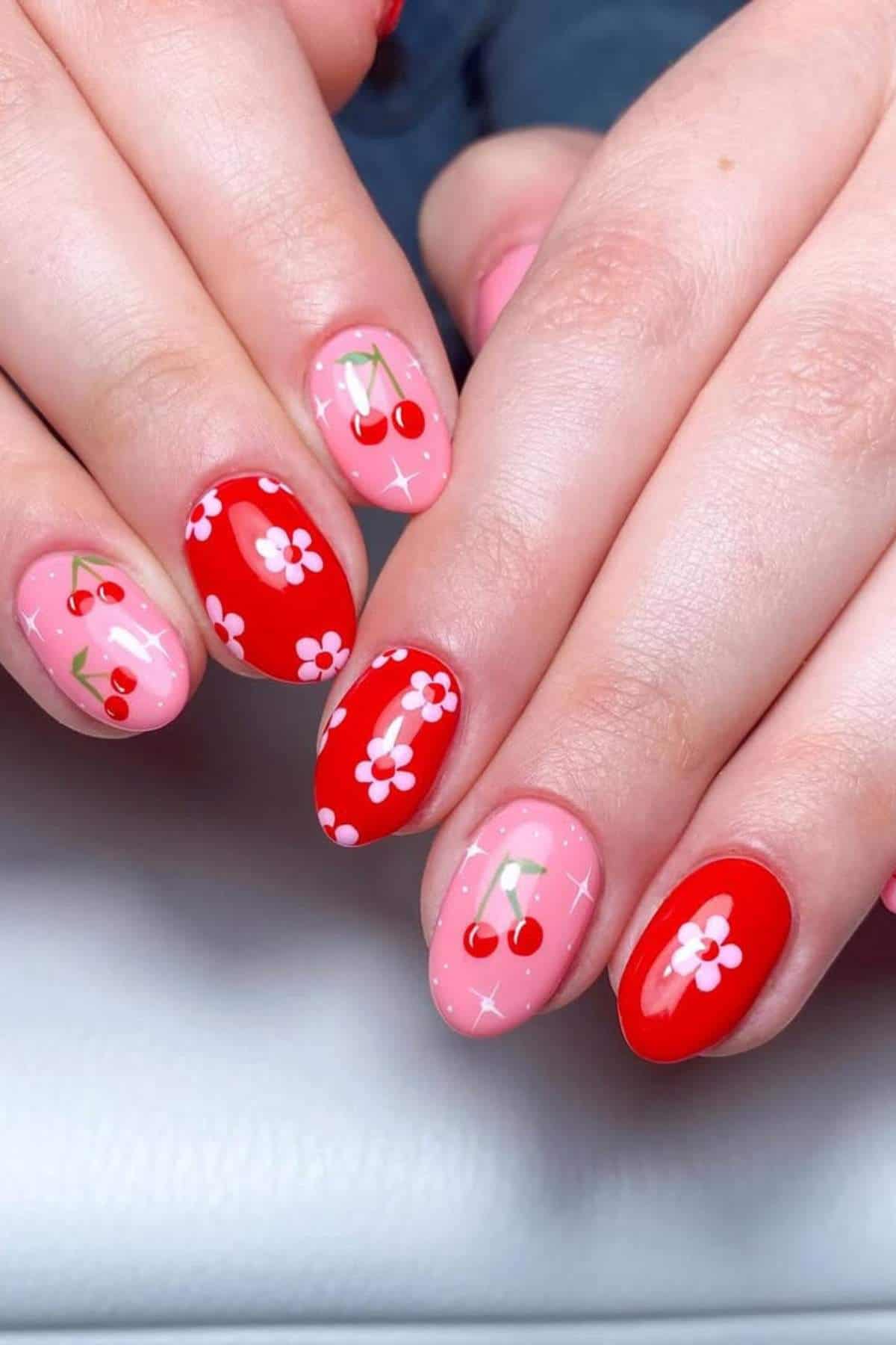 Hands with manicured nails featuring pink and red polish, decorated with cherry and white flower designs.