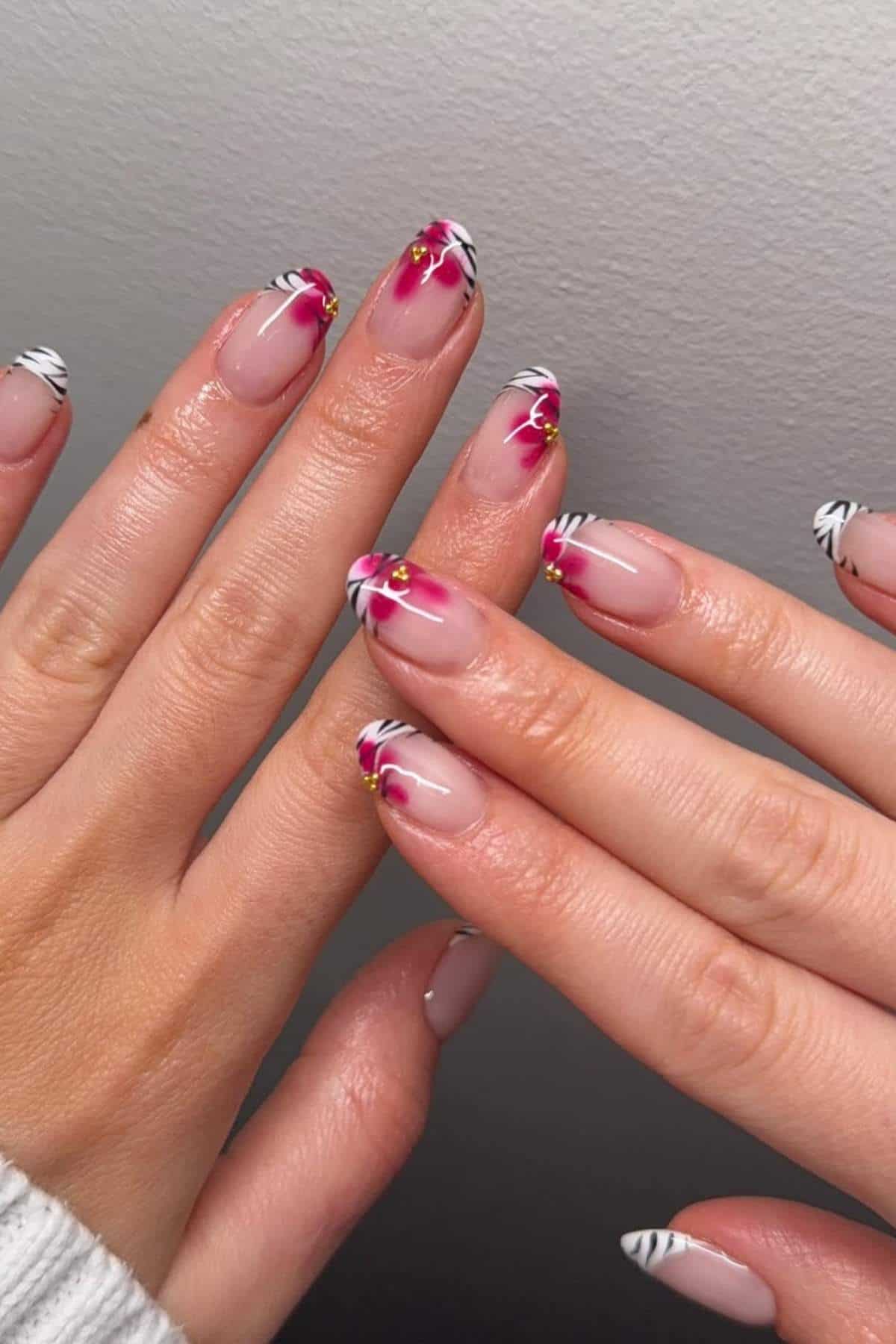 Hands with medium-length nails featuring a pink, white, and black zebra-stripe and floral nail art design with gold accents.