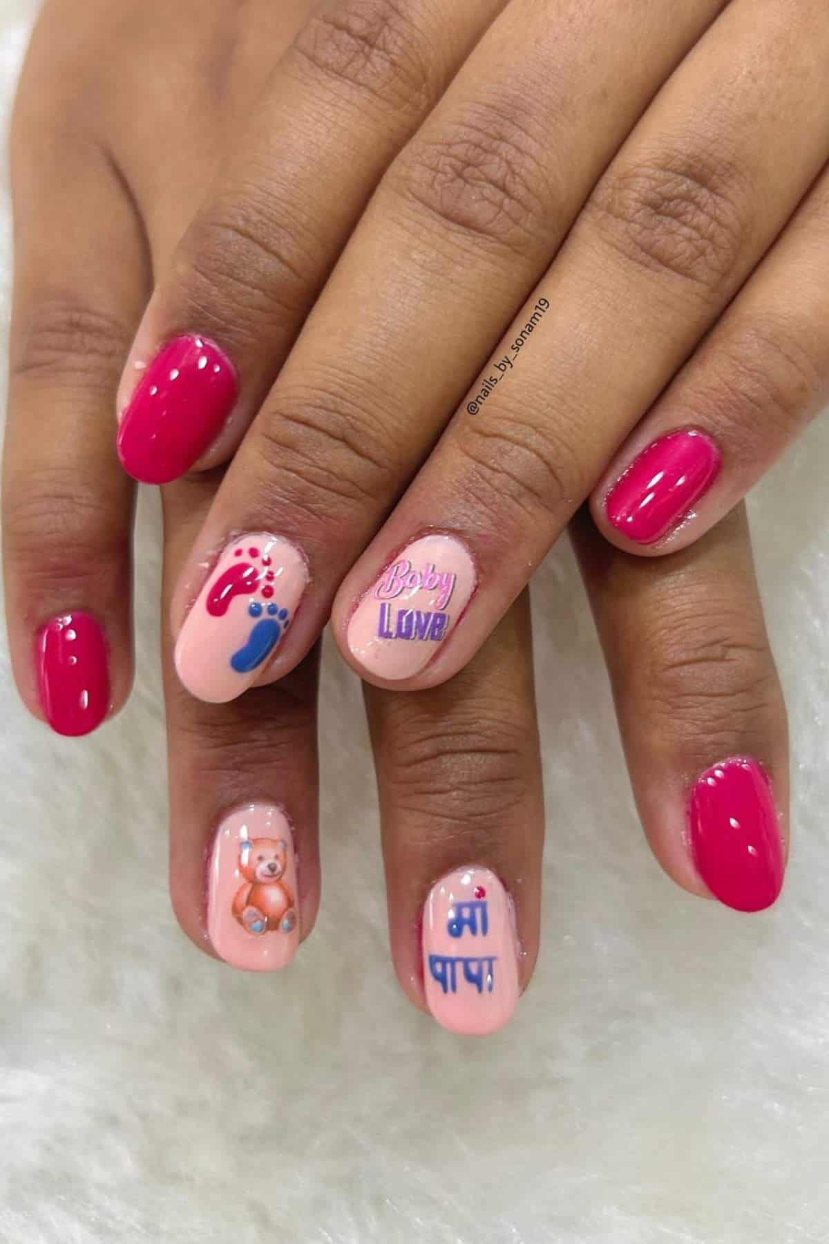 Hands with pink and nude manicure; accent nails feature baby-themed designs, including tiny footprints, a teddy bear, "Baby Love," and text in Hindi.