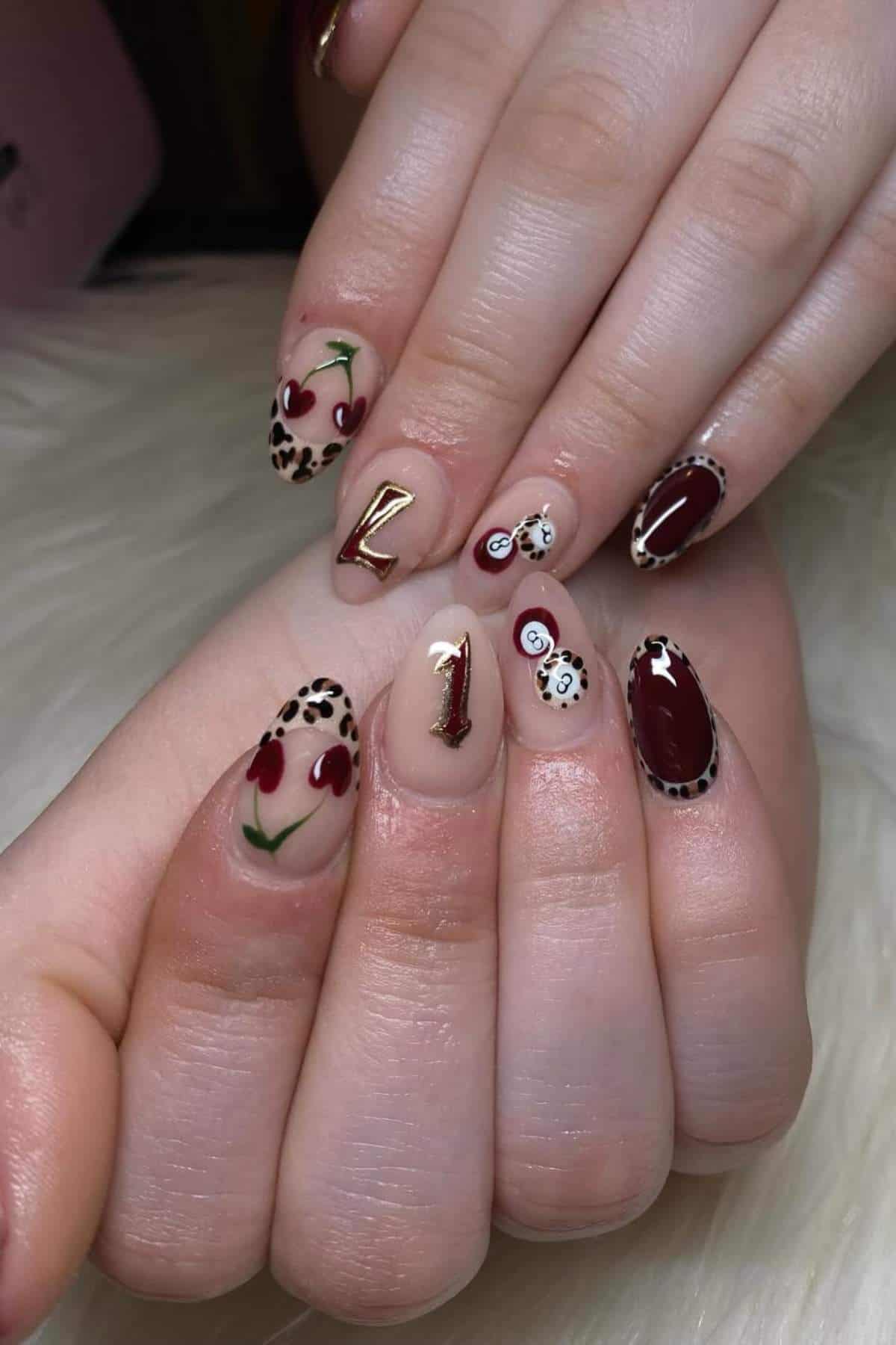 Hands with almond-shaped nails featuring nail art designs: cherries, leopard print, gold numbers, red accents, and white cartoon faces on a light background.