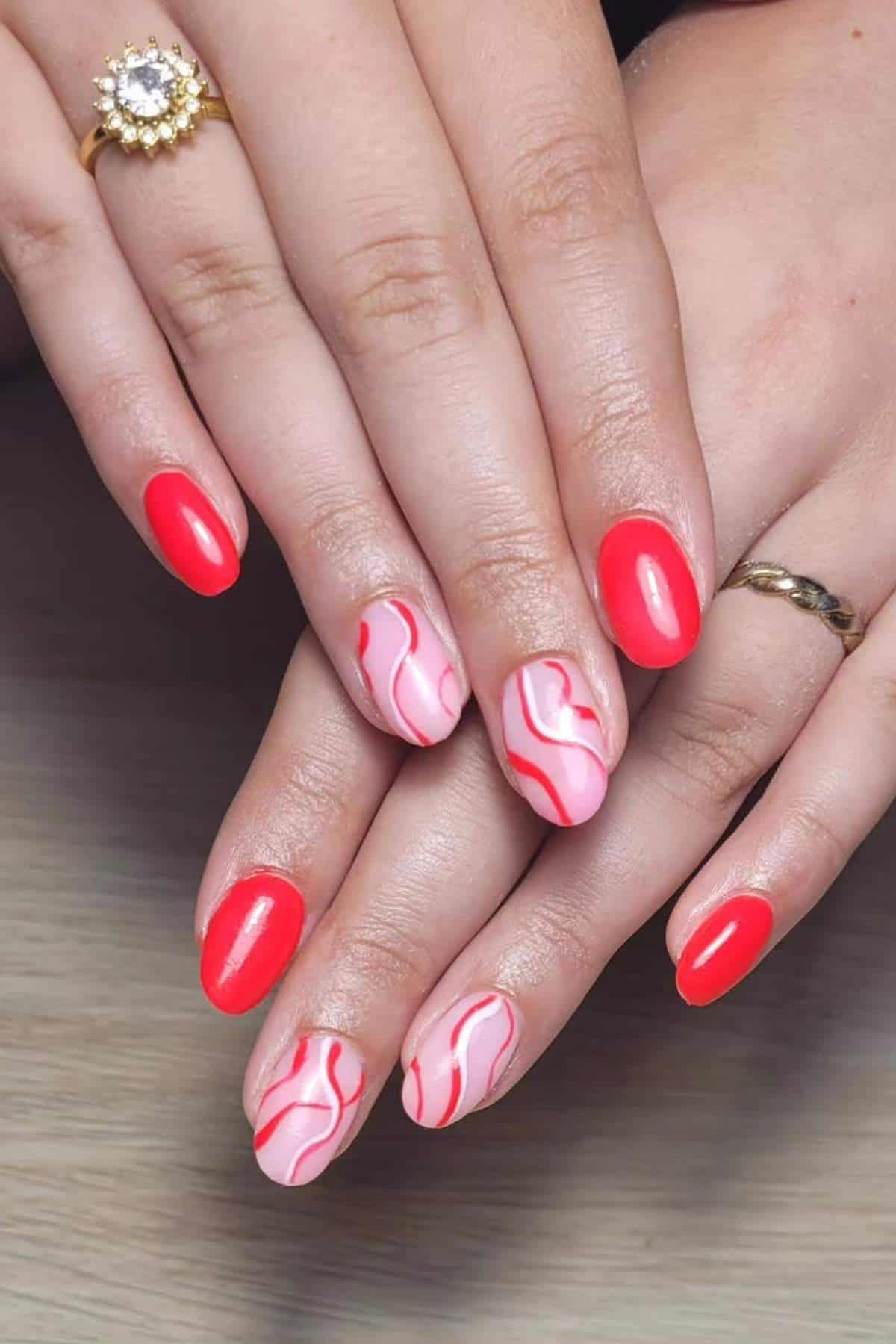 Hands with short, oval-shaped nails painted in glossy red and pink with wavy red line designs on two accent nails. Two gold rings are worn on the fingers.