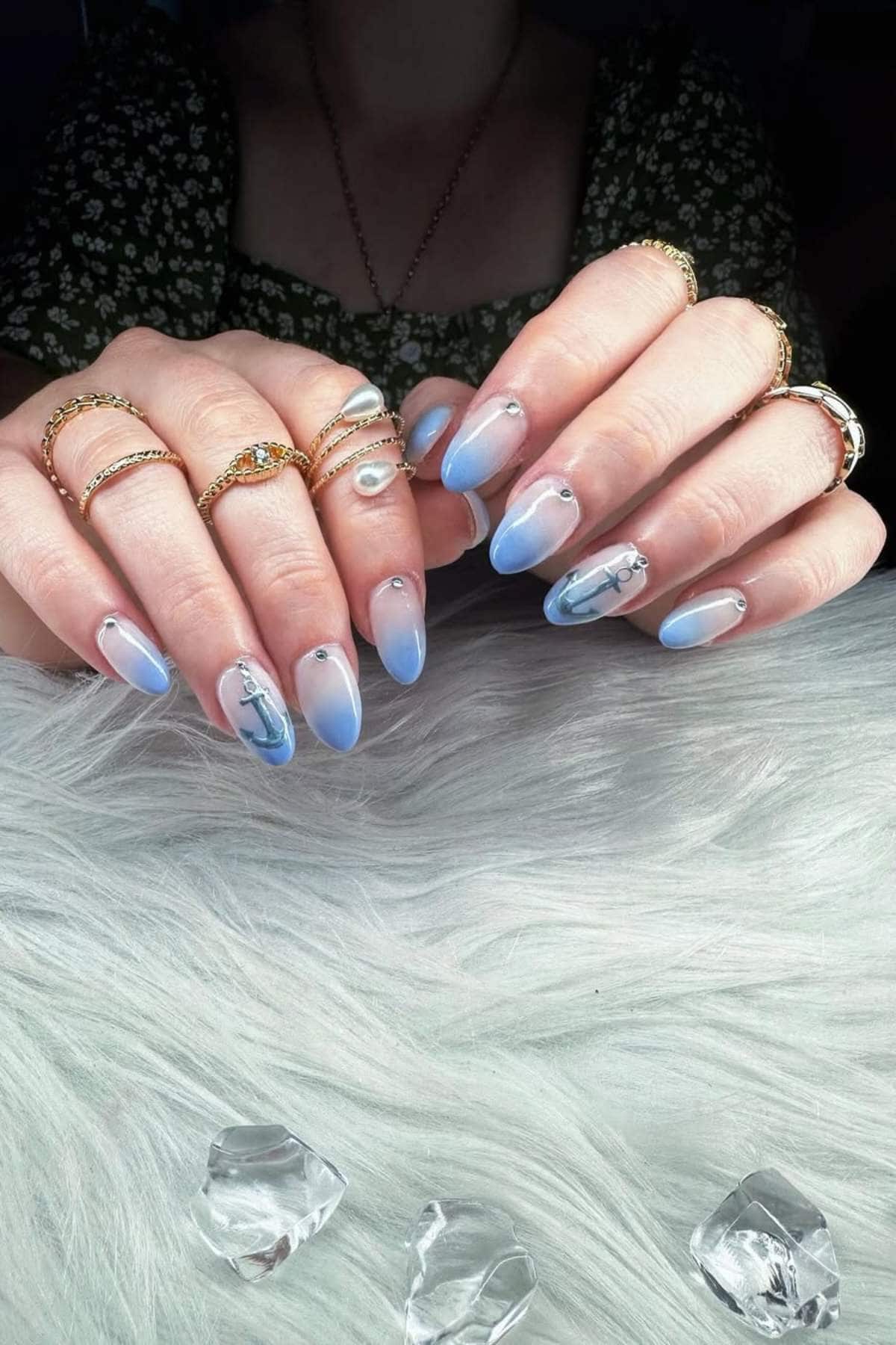 Hands with long, almond-shaped nails painted in light blue and white gradient, featuring small rhinestones and nautical-themed nail art, resting on a white fur surface with clear crystals.