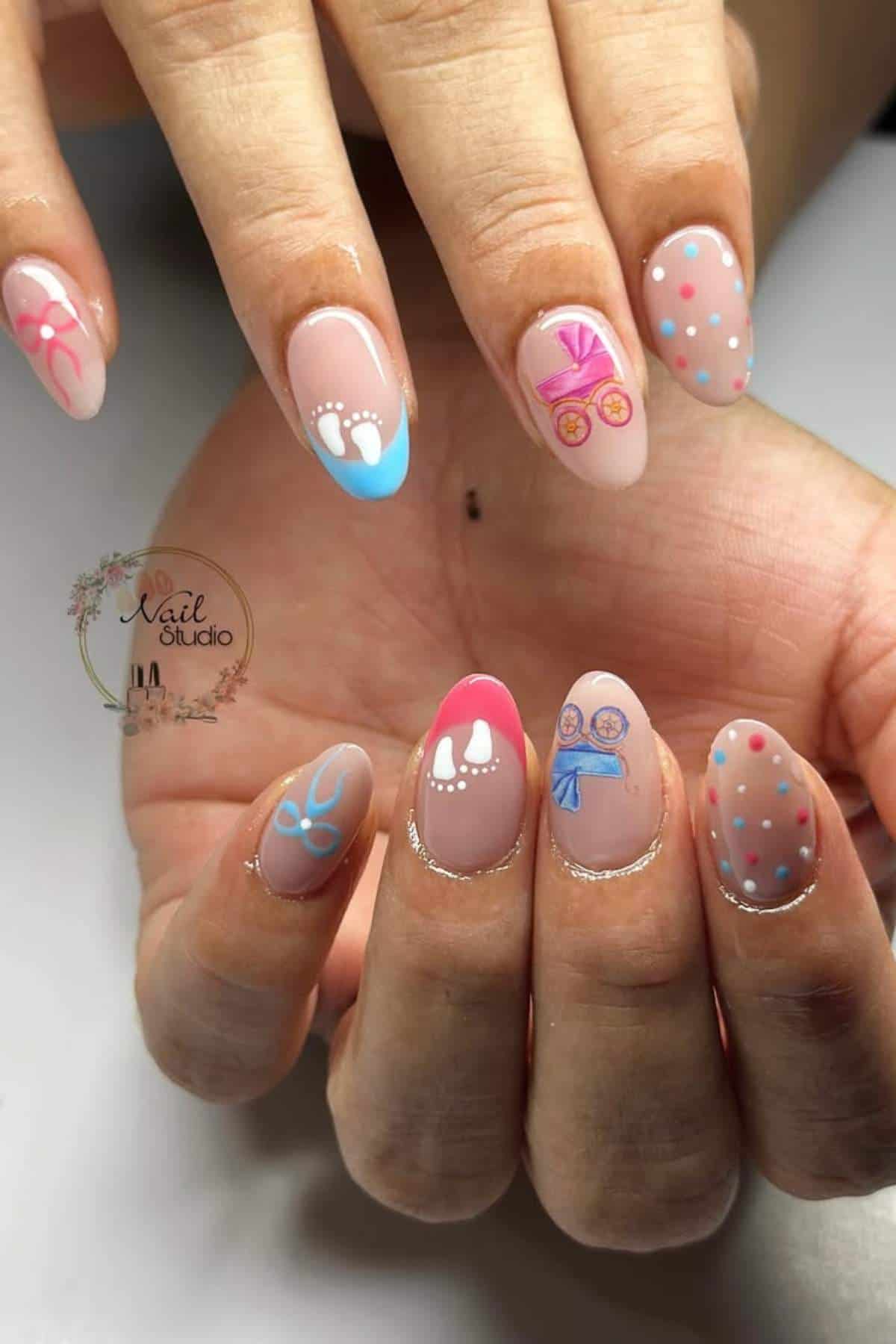 Hands with almond-shaped nails featuring baby-themed nail art, including baby feet, stroller, bib, pacifier, and blue and pink polka dots on a nude background.