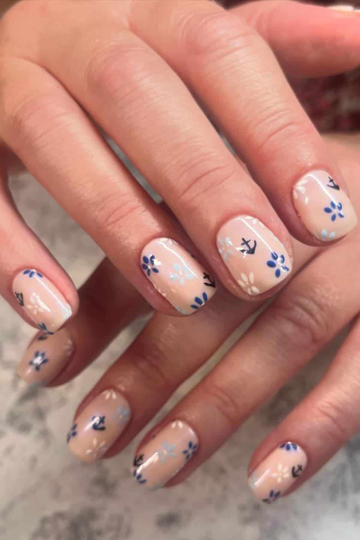 Close-up of hands with nude-colored nail polish featuring small blue and white floral designs on each fingernail.