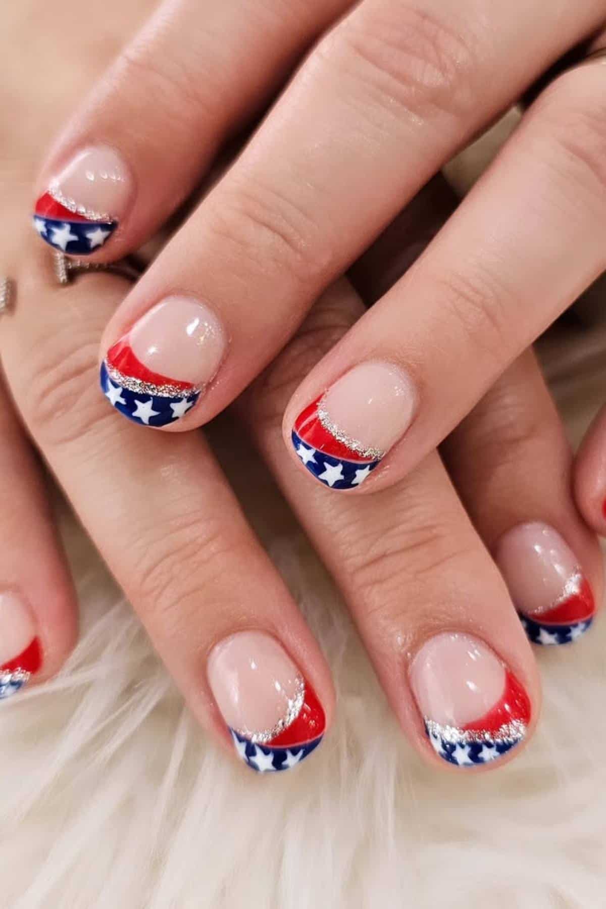 Close-up of hands with nails painted in a red, white, and blue French tip design featuring white stars and silver glitter accents.