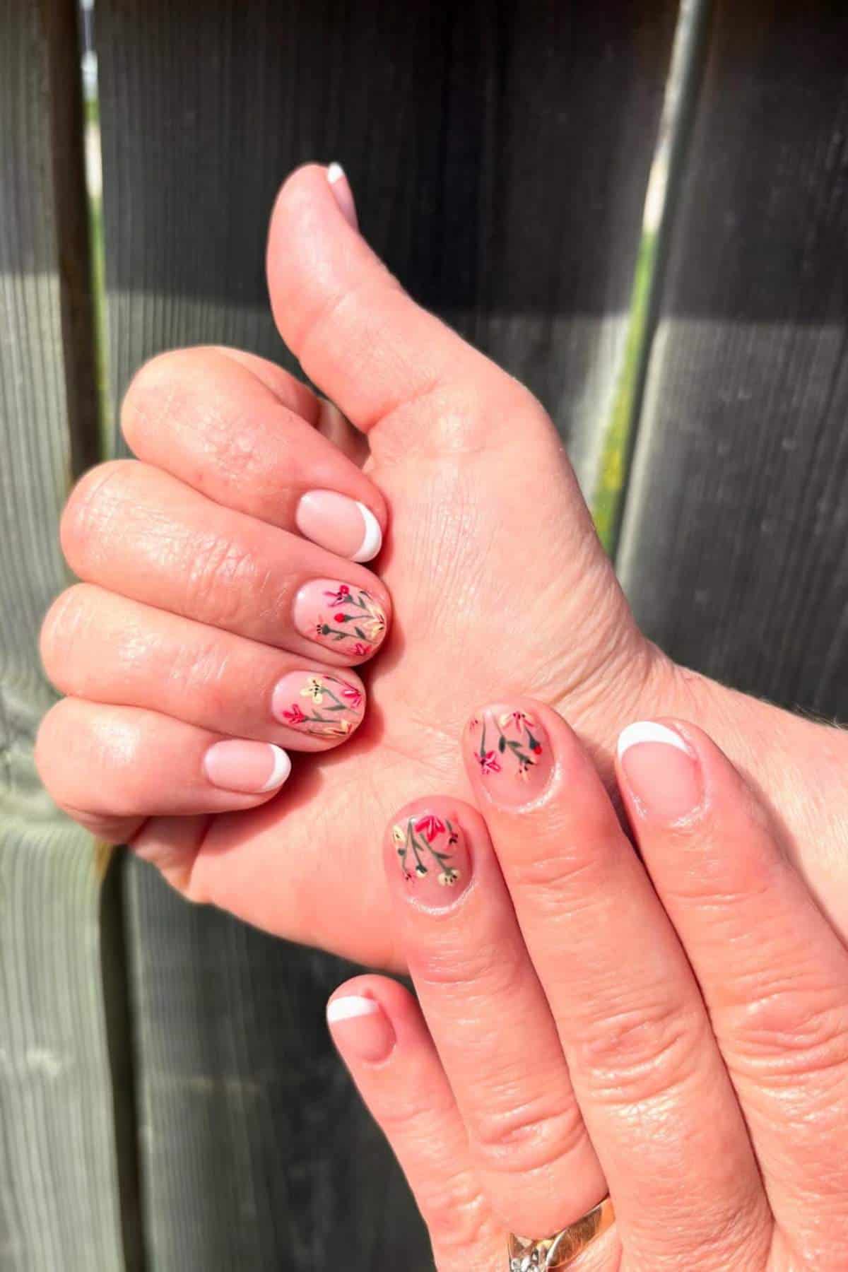 A hand with short nails featuring a French manicure and floral nail art on two fingers, held against a wooden background.
