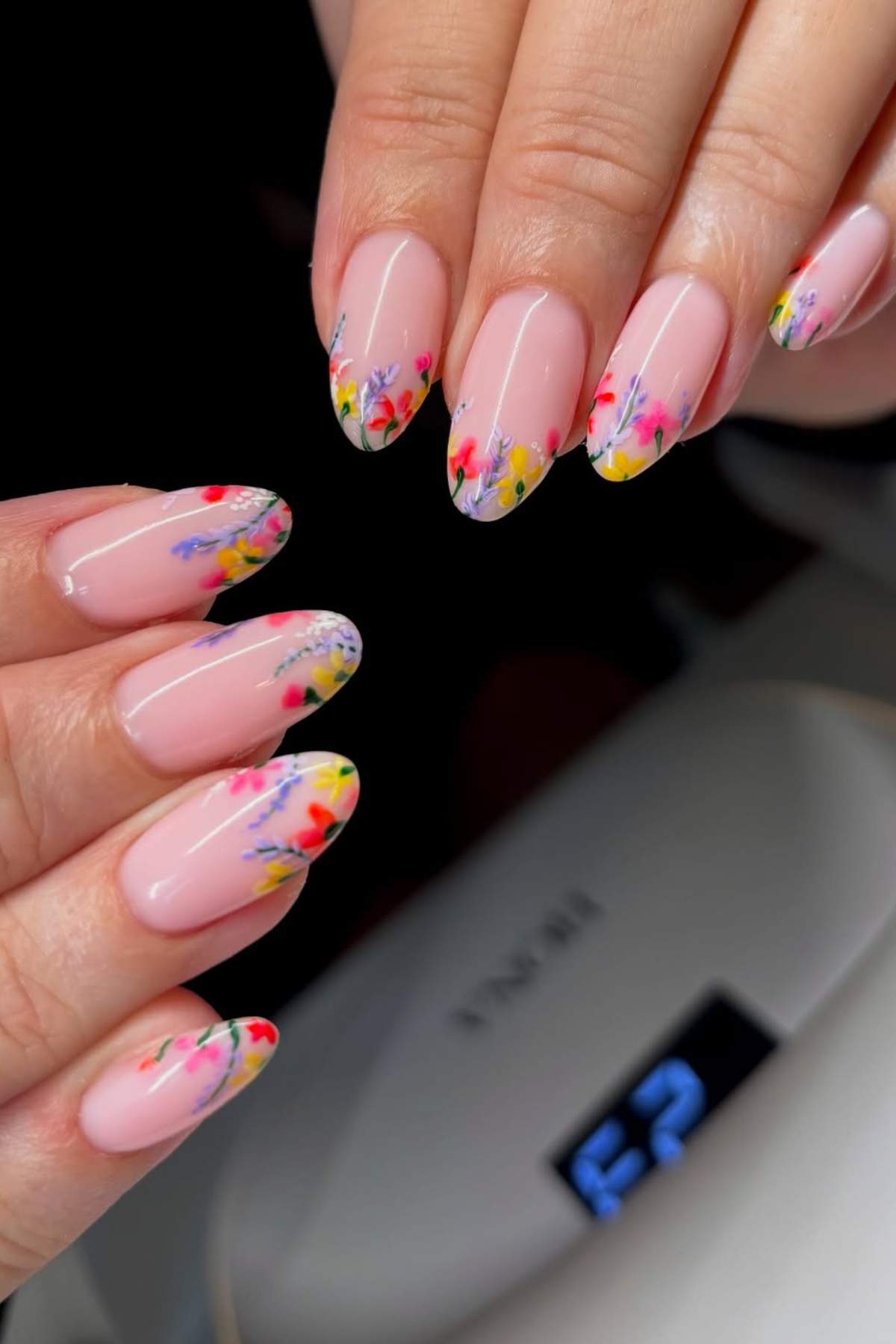 Close-up of hands with almond-shaped, nude-pink nails featuring colorful floral designs on the tips.