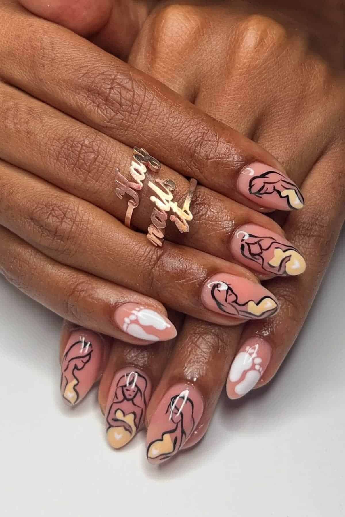 A close-up of hands with almond-shaped nails painted with abstract line art of female figures in nude, white, and black tones. The person also wears a gold ring with cursive text.