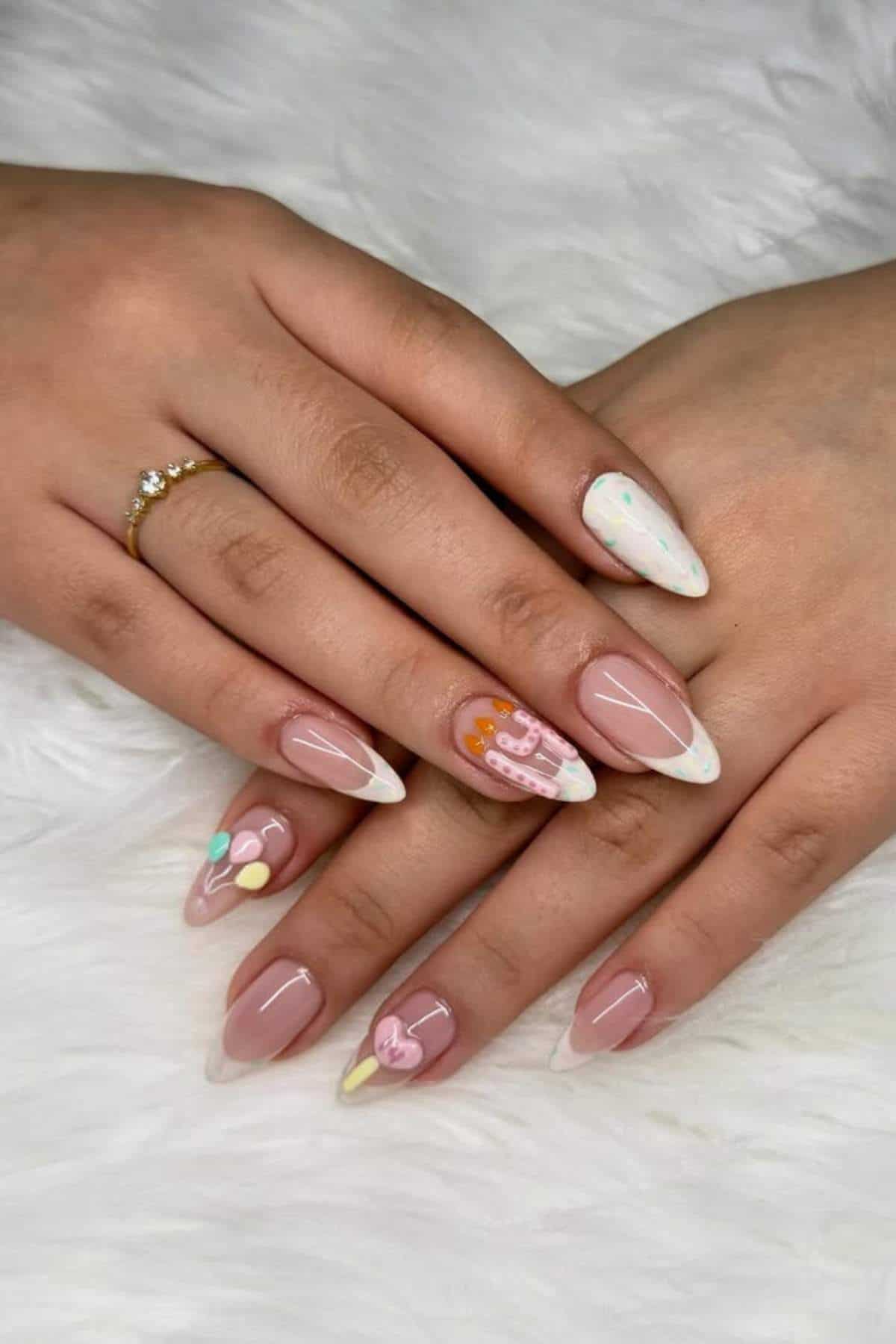 Hands with almond-shaped nails featuring pastel-colored abstract designs and a beaded ring on one finger, resting on a white textured surface.