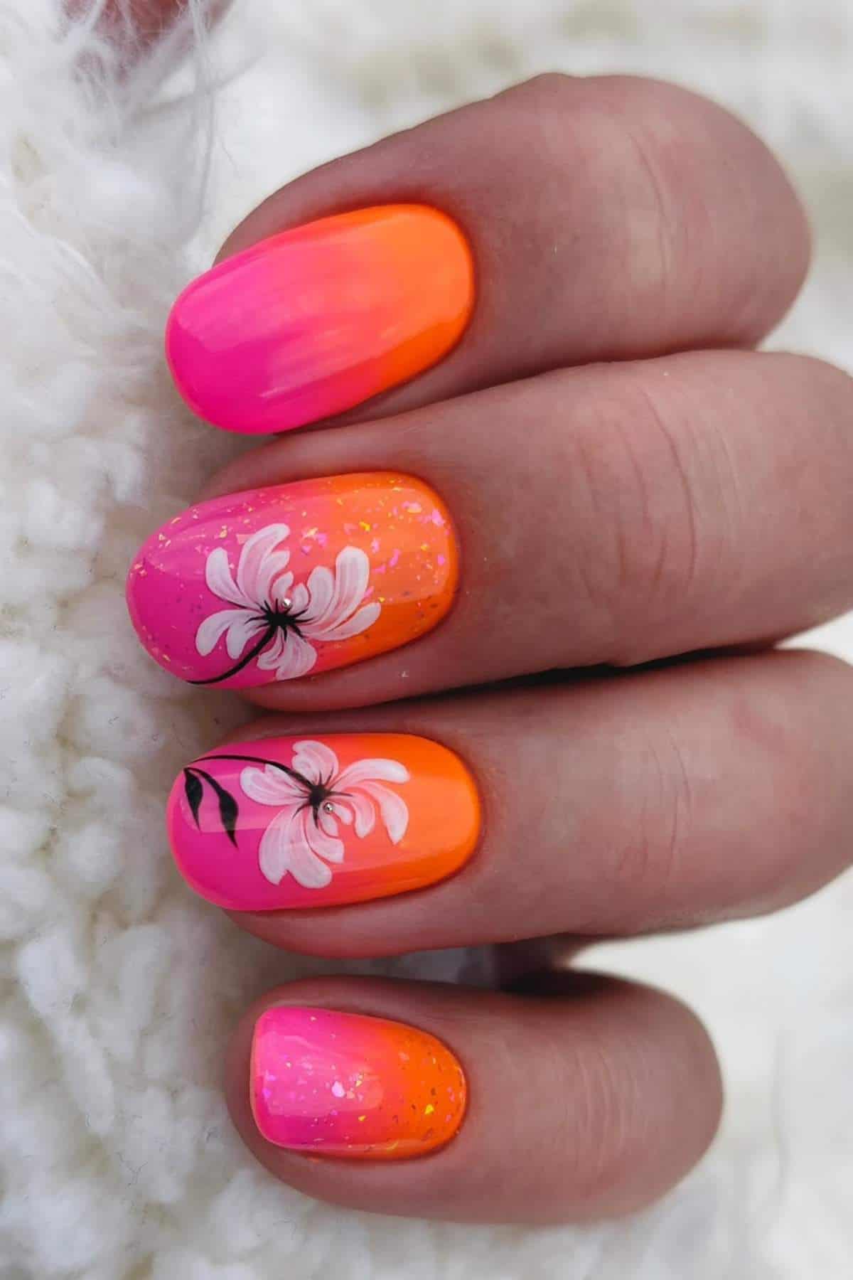 A hand with bright pink and orange ombre nails, two nails featuring white floral designs and delicate black accents, against a soft white textured background.