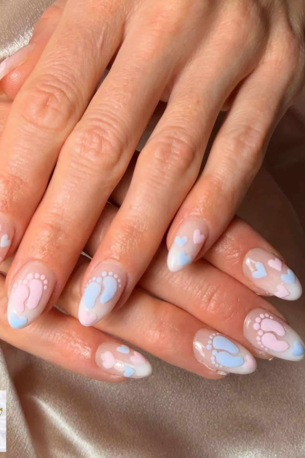 Close-up of hands with manicured nails featuring blue and pink baby footprints and heart designs on a pale background.