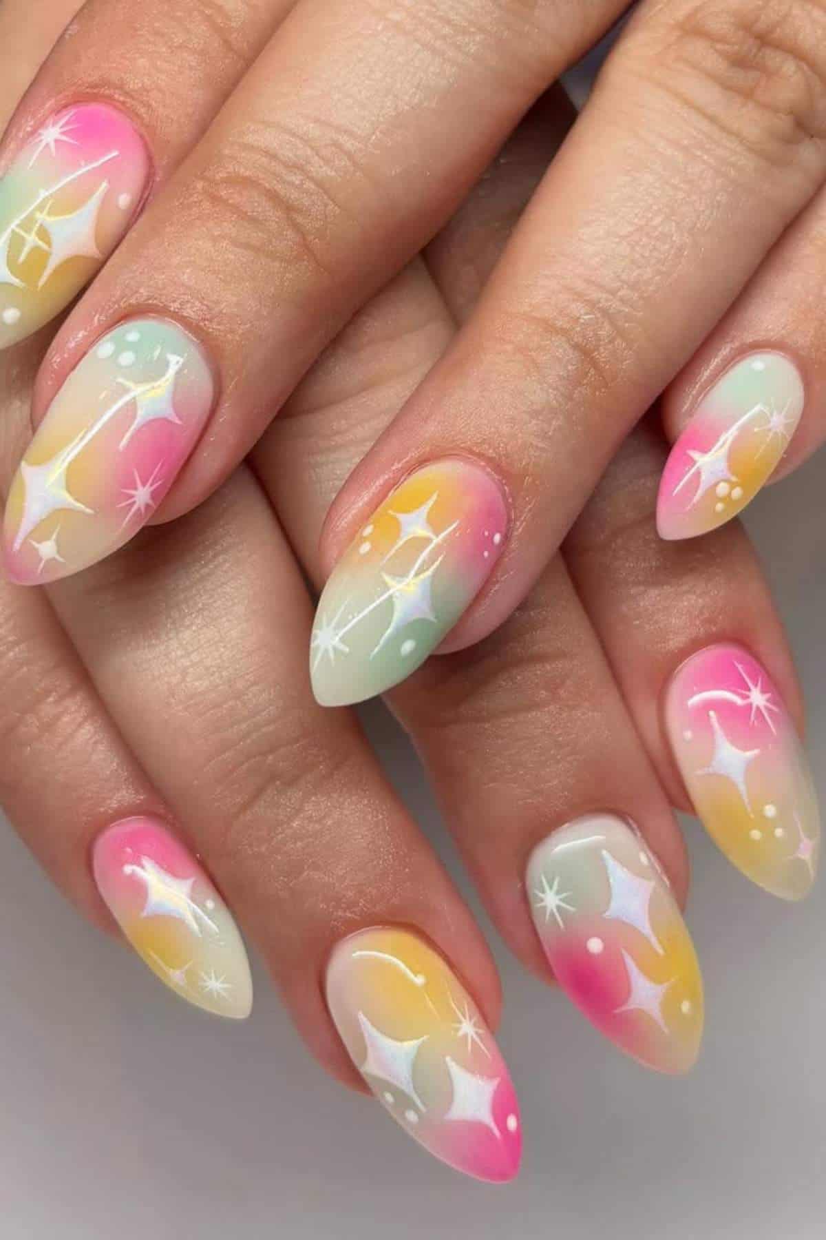 Hands with almond-shaped nails painted in pastel pink, yellow, and mint gradients, decorated with white star and dot designs.