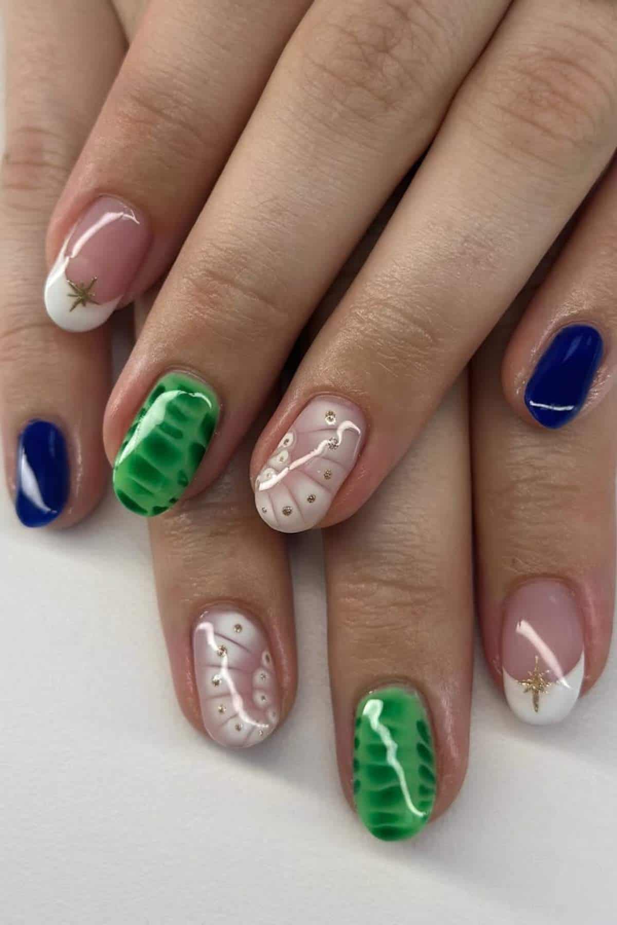 Close-up of hands with manicured nails featuring mixed designs: blue, green textured, white tips with gold stars, and intricate white patterns with small gold accents.