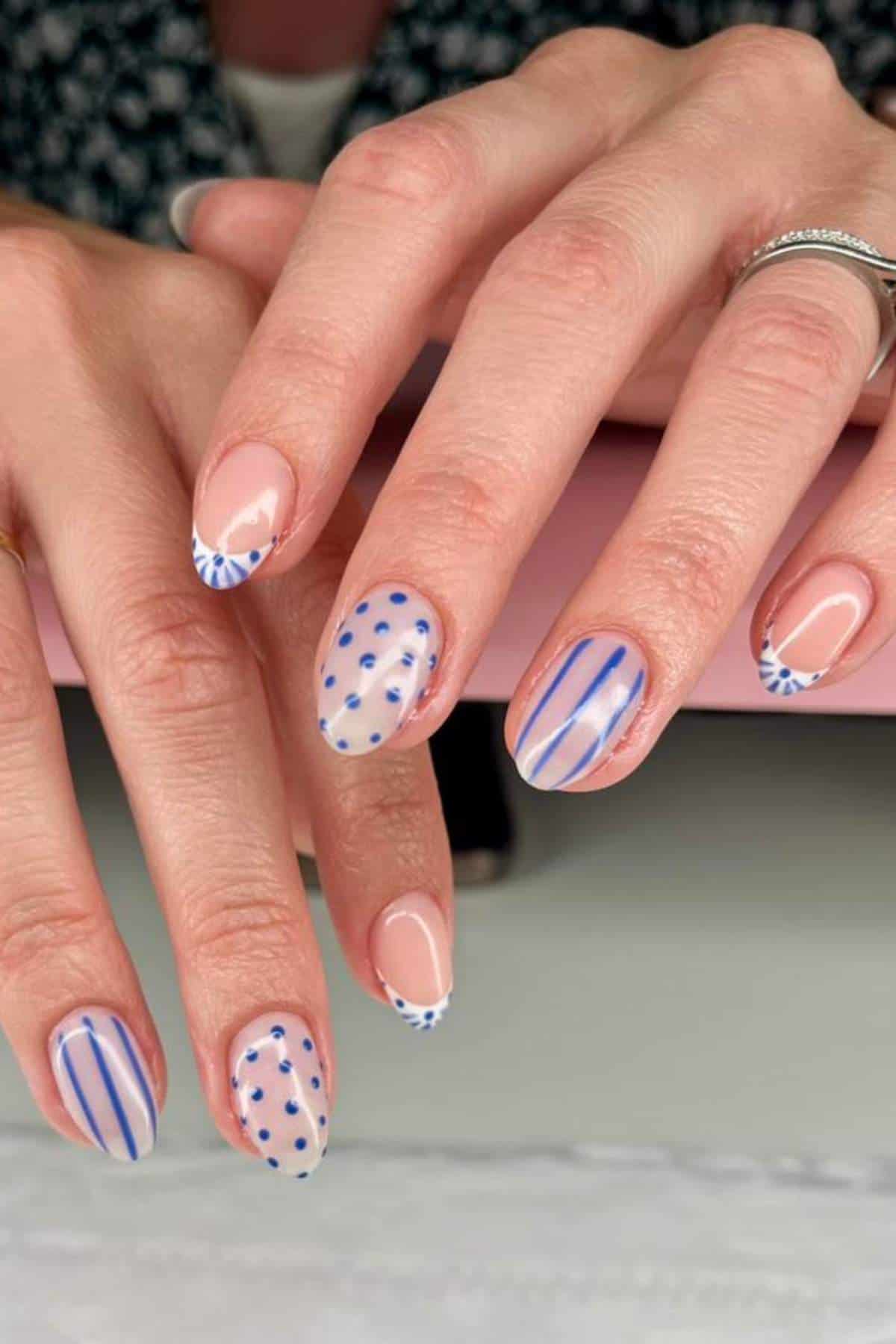 Close-up of hands with nude nail polish featuring blue polka dots, stripes, and geometric tip designs. The person is wearing silver rings.