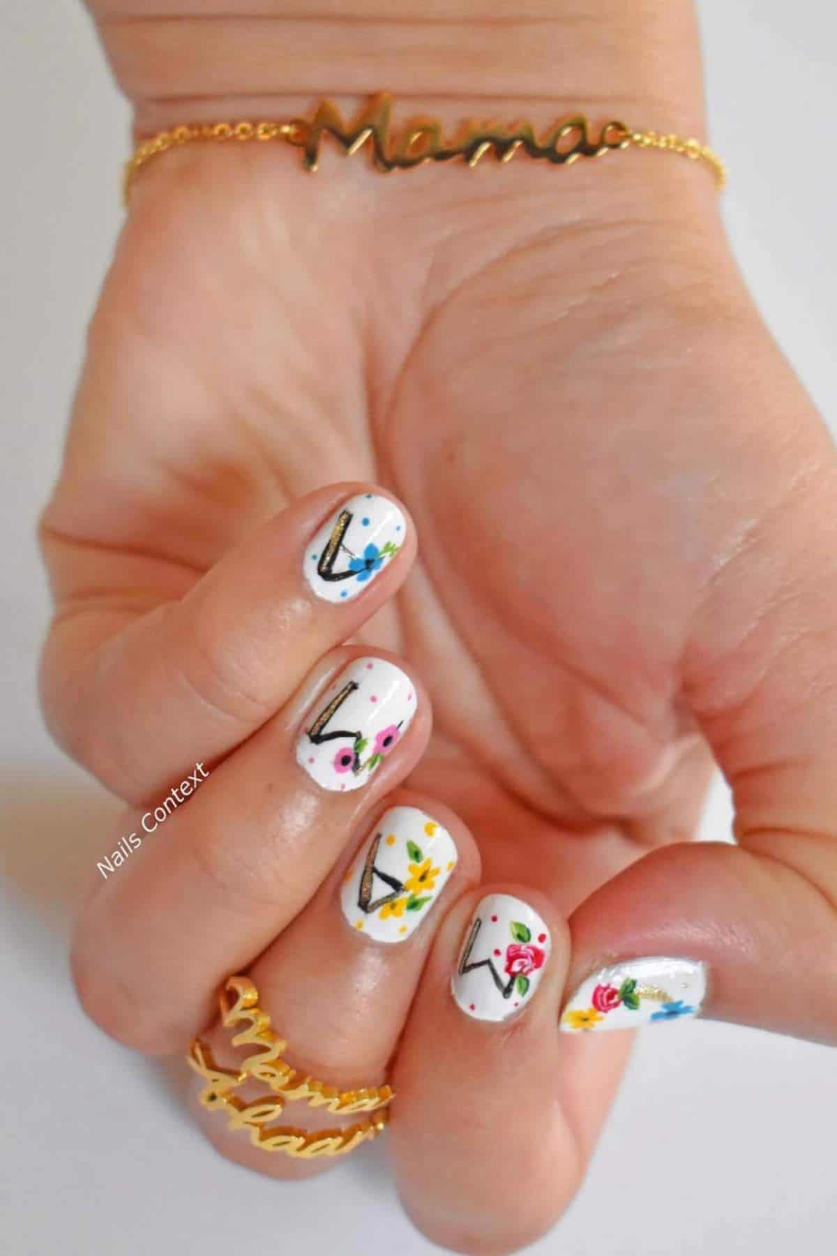 A hand with "Mama" jewelry displays white nails decorated with colorful, abstract floral and line art designs.