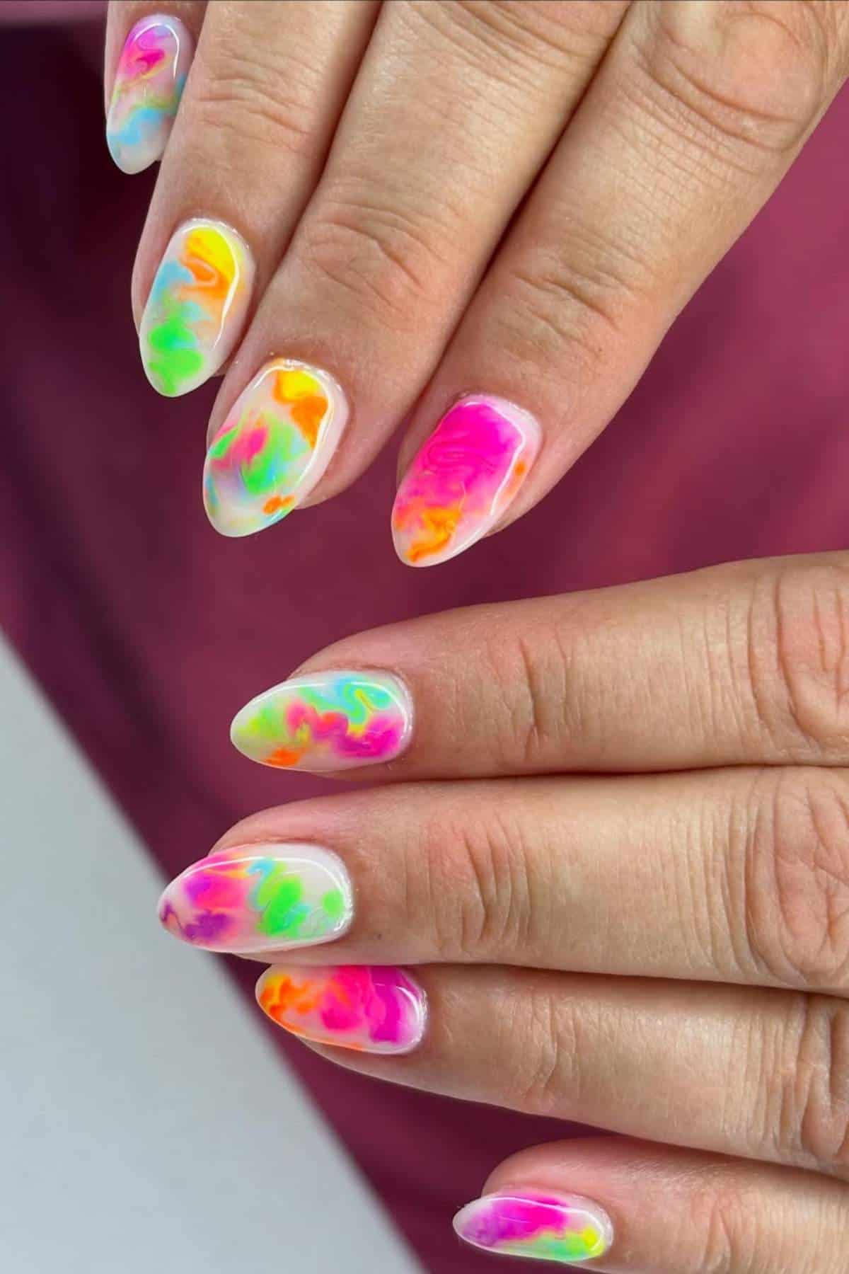 Close-up of hands with almond-shaped nails featuring a bright, multicolored watercolor marble nail art design on a white base.
