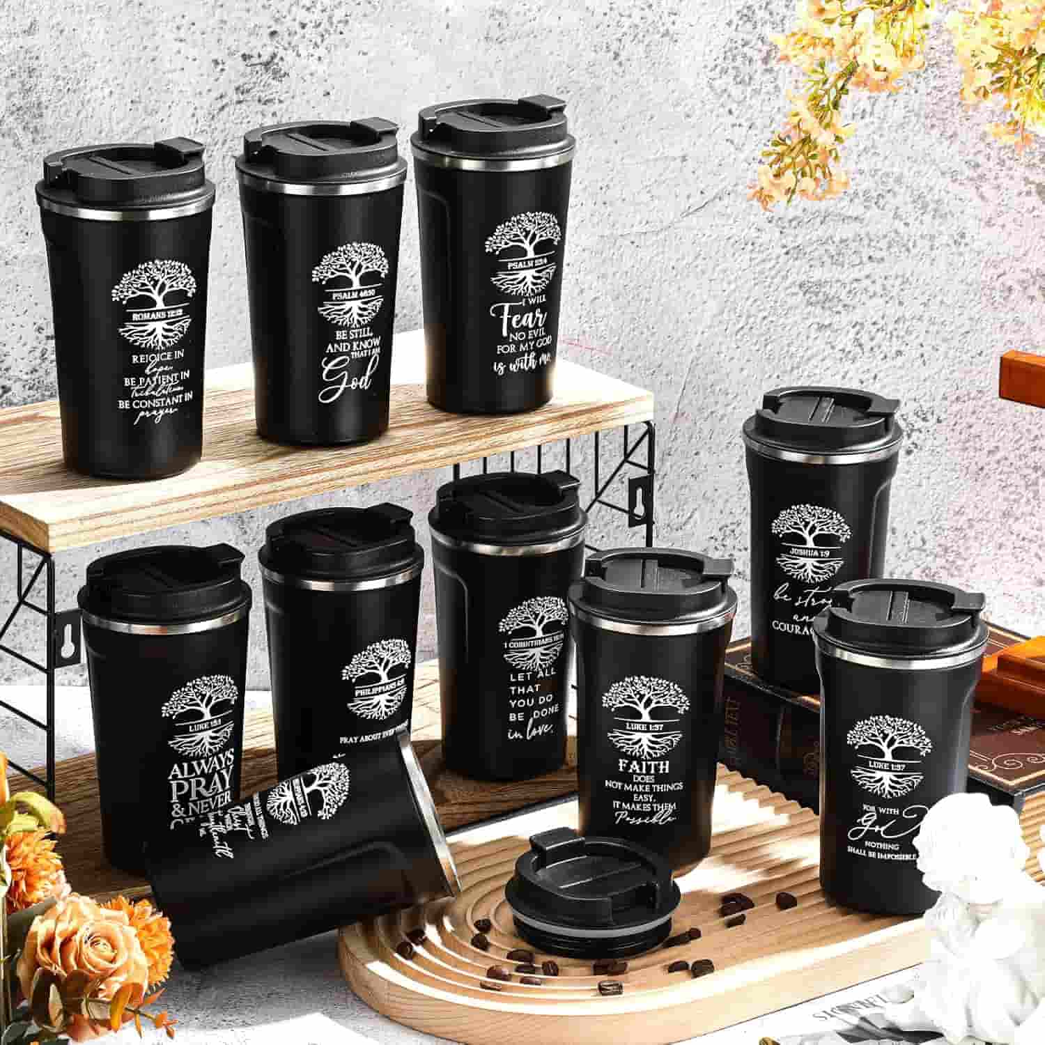 Nine black travel mugs with inspirational quotes and tree designs are arranged on a table with scattered coffee beans and a wooden tray, against a textured background.
