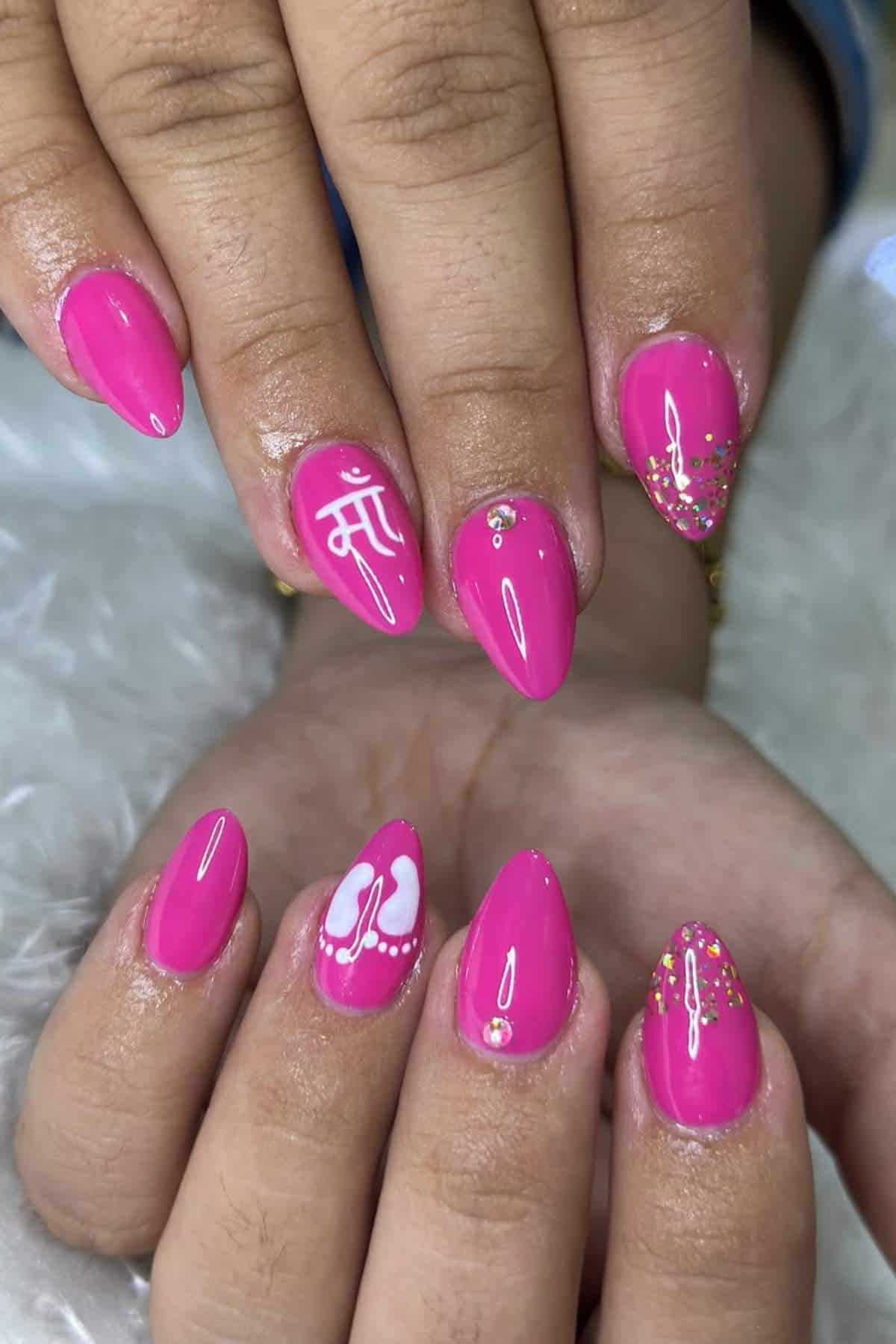 Close-up of hands with bright pink almond-shaped nail art, featuring white designs, gold glitter, and small rhinestones.