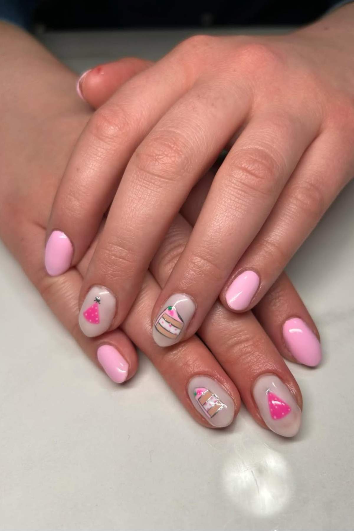 Hands with light pink nail polish; some nails feature birthday-themed designs, including a cake and party hat illustrations on a neutral background.