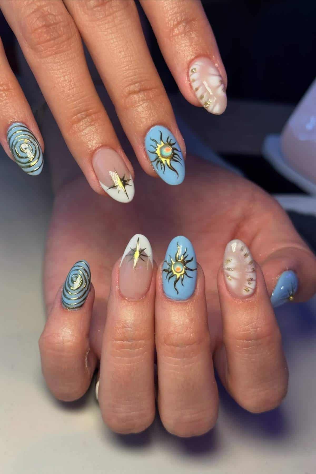 A hand with almond-shaped nails features blue, white, and nude polish with designs of suns, eyes, and dots in gold, black, and white accents.