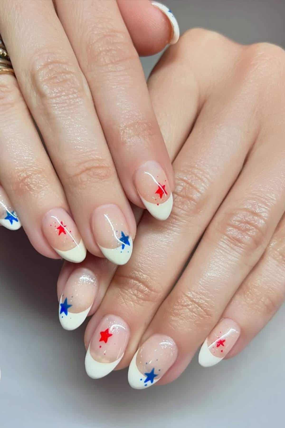 Hands with almond-shaped nails featuring white French tips and red and blue star decorations on a neutral base.