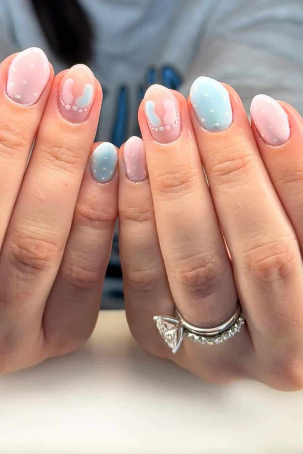 Hands with pastel nail art featuring blue, pink, and white abstract designs, small dots, and a silver ring with a large triangular stone.