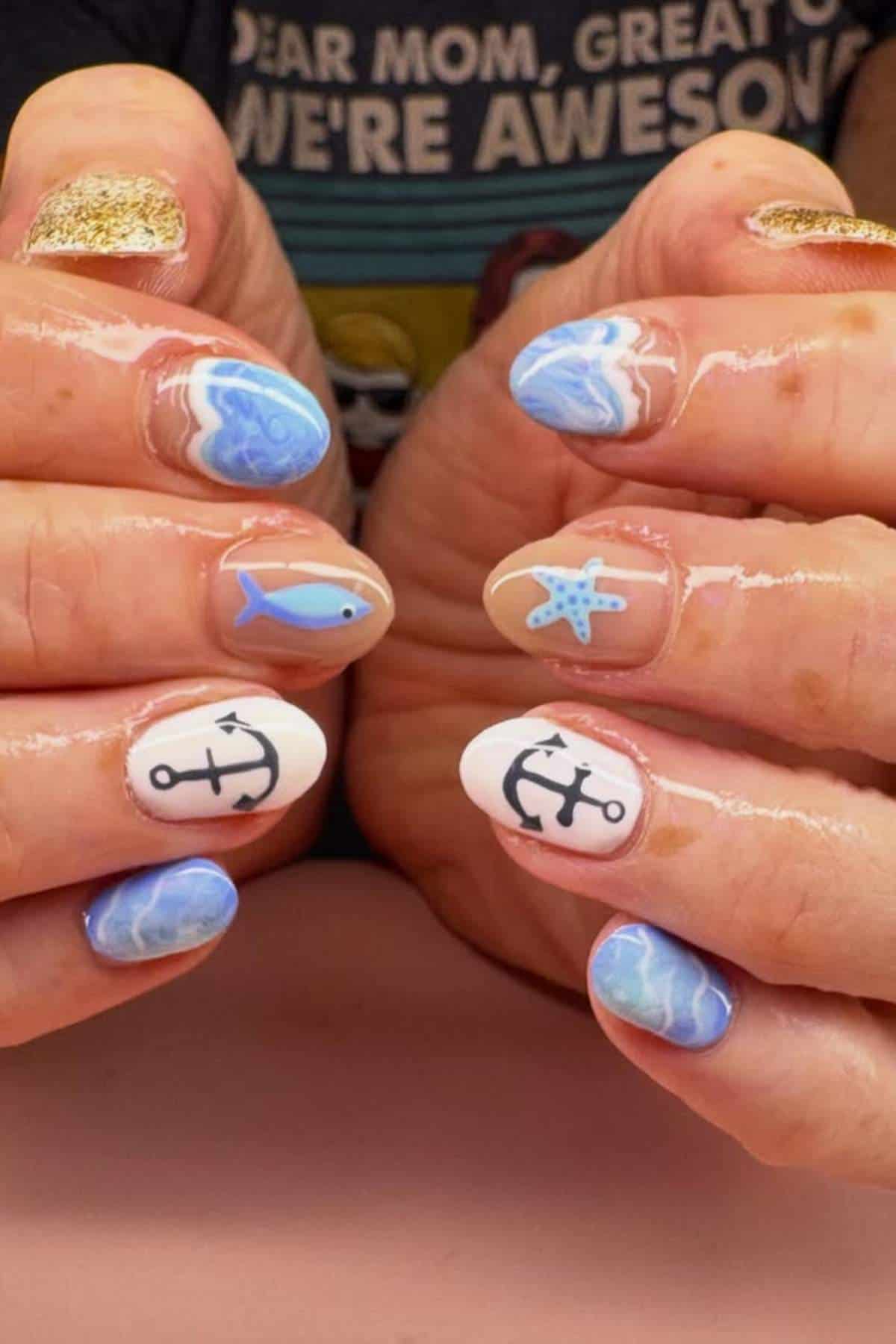Hands with nautical-themed nail art featuring anchors, a fish, a starfish, ocean waves, and gold glitter on some nails.