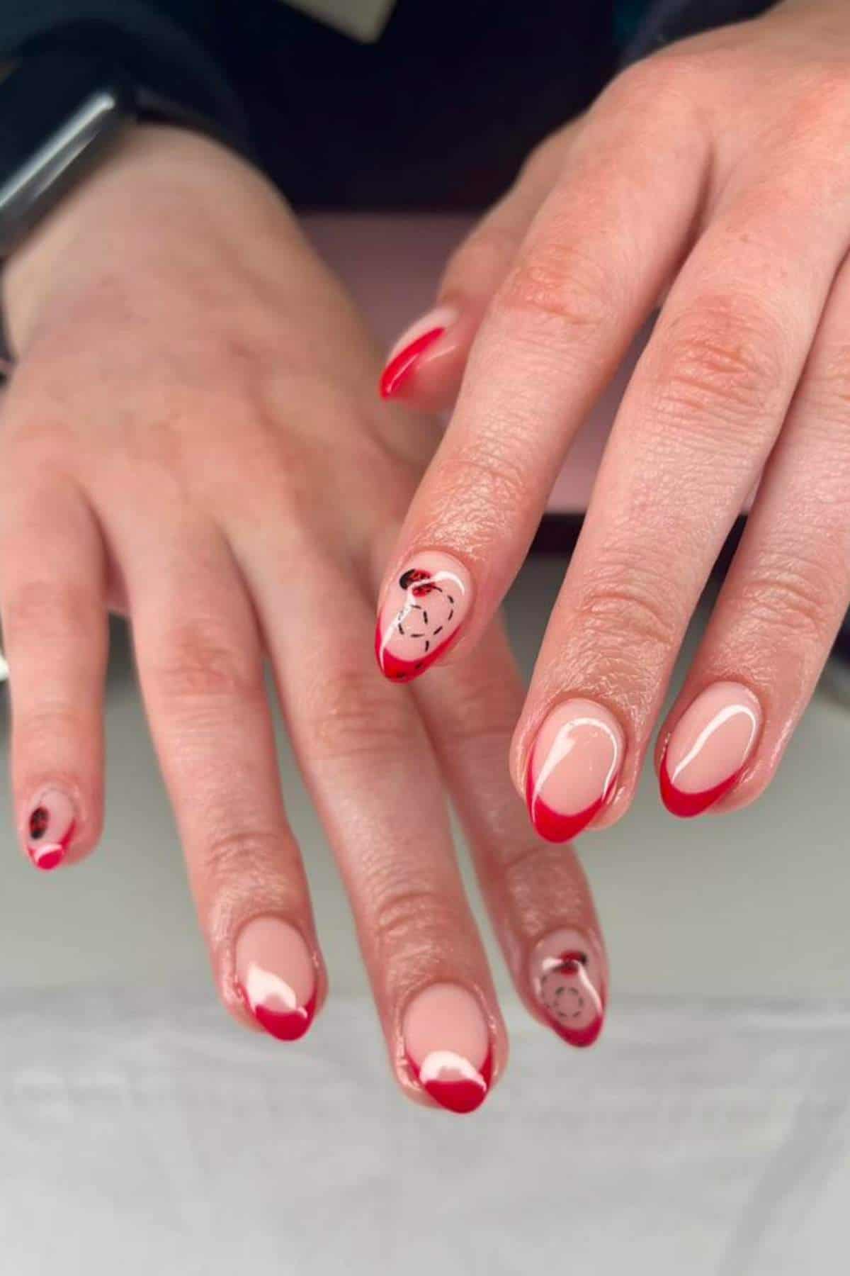 Hands with almond-shaped nails featuring a nude base, red French tips, and delicate black abstract line and dot designs on a few accent nails.
