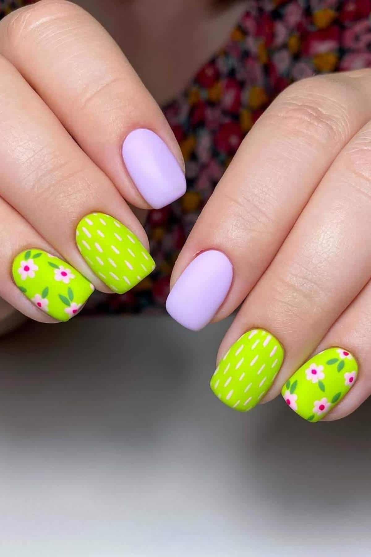 Close-up of hands with manicured nails featuring pastel purple and green polish; green nails have floral and striped designs.